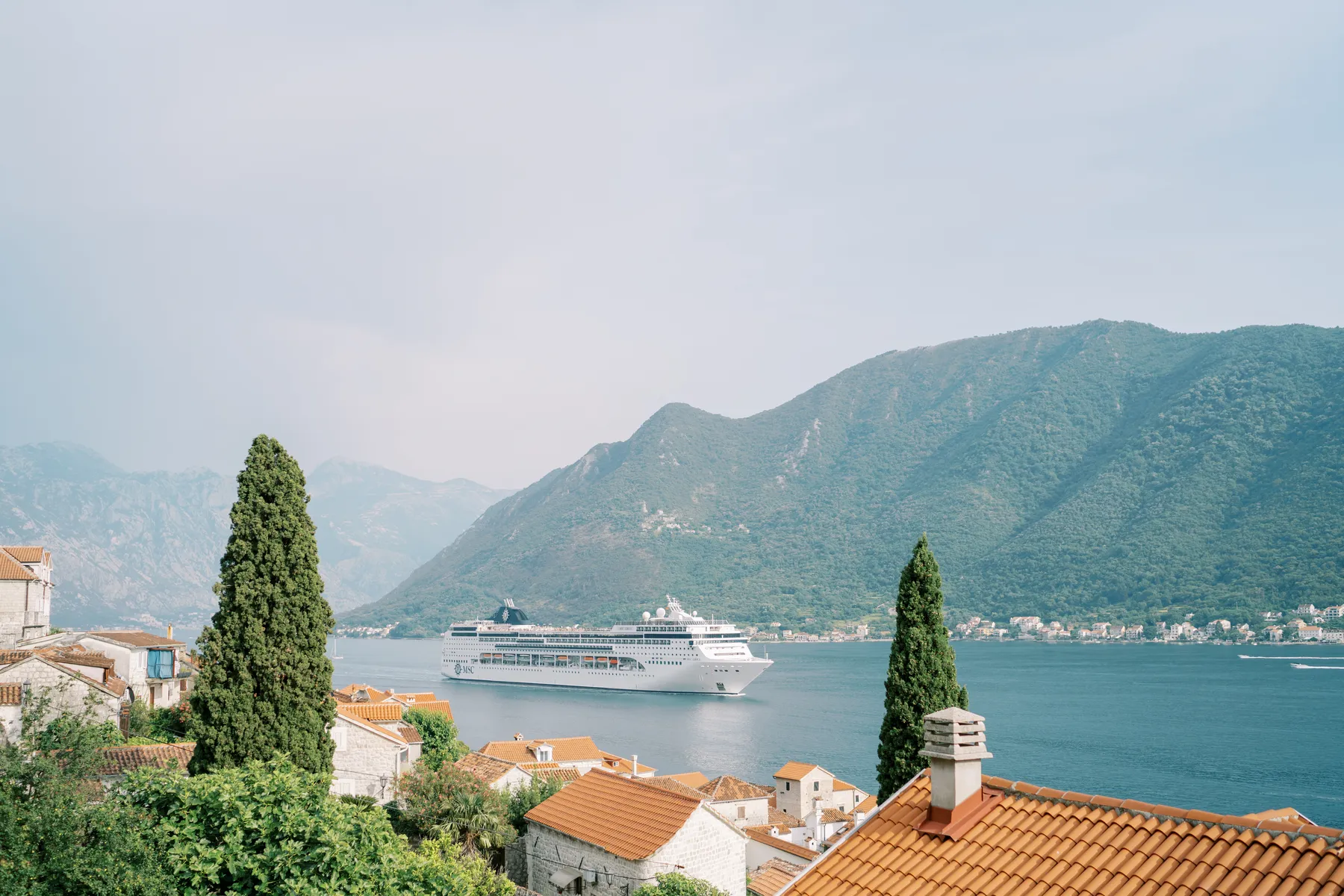 A white cruise ship sails through the Bay of Kotor past terracotta rooftops and cypress trees in Perast, Montenegro.