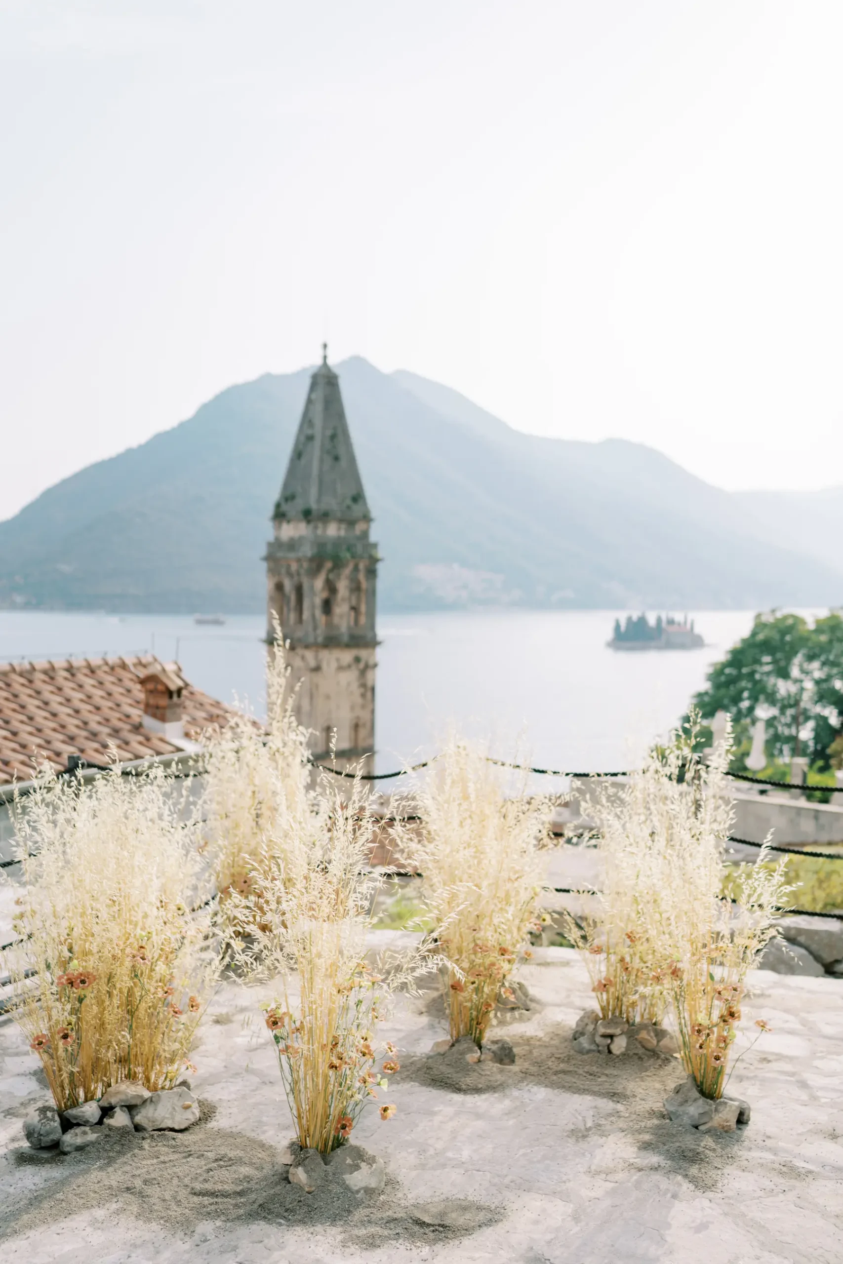 Floral grass arrangements on a terrace overlook a church steeple, bay and mountain in Perast, Montenegro.