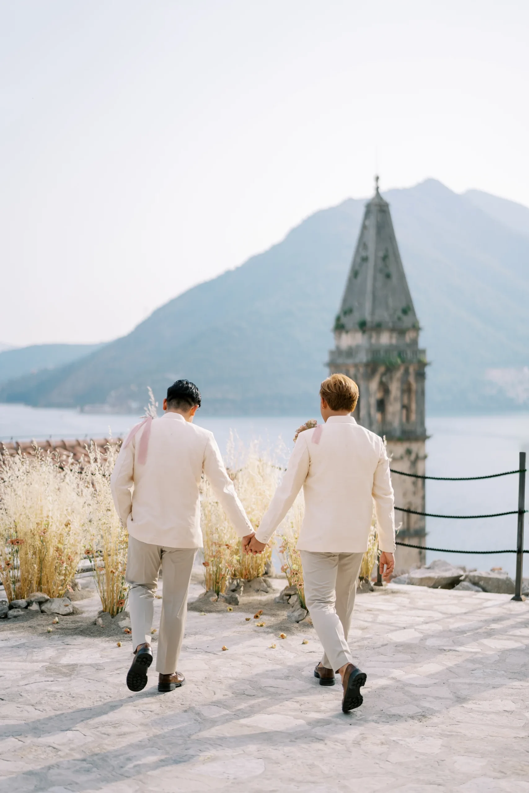 Ryan Jake Pisueña and Jori Blas Lazalita hold hands walking toward a stone church tower in Perast, Montenegro.