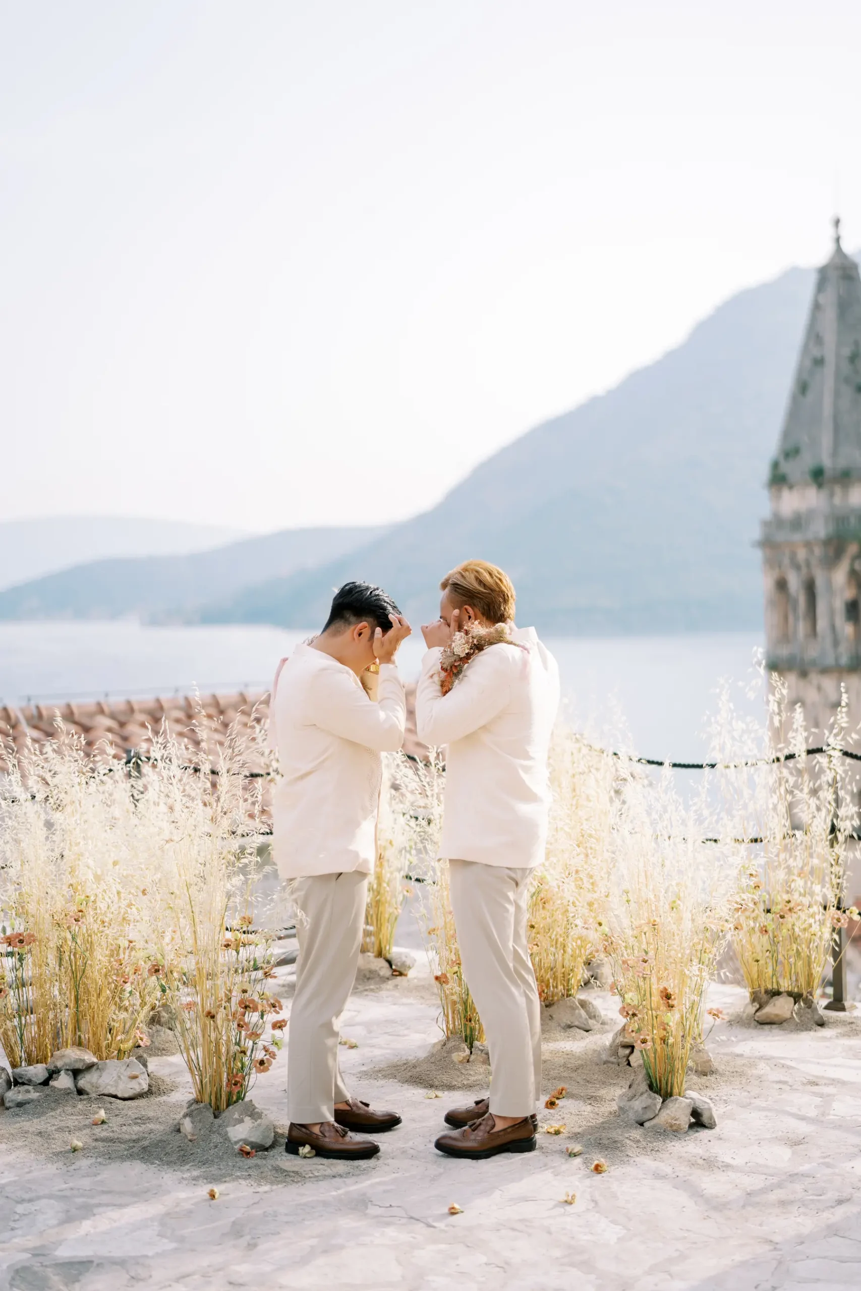Ryan Jake and Jori face each other in white suits amid wildflowers at Monte Bay Retreat in Perast, Montenegro.