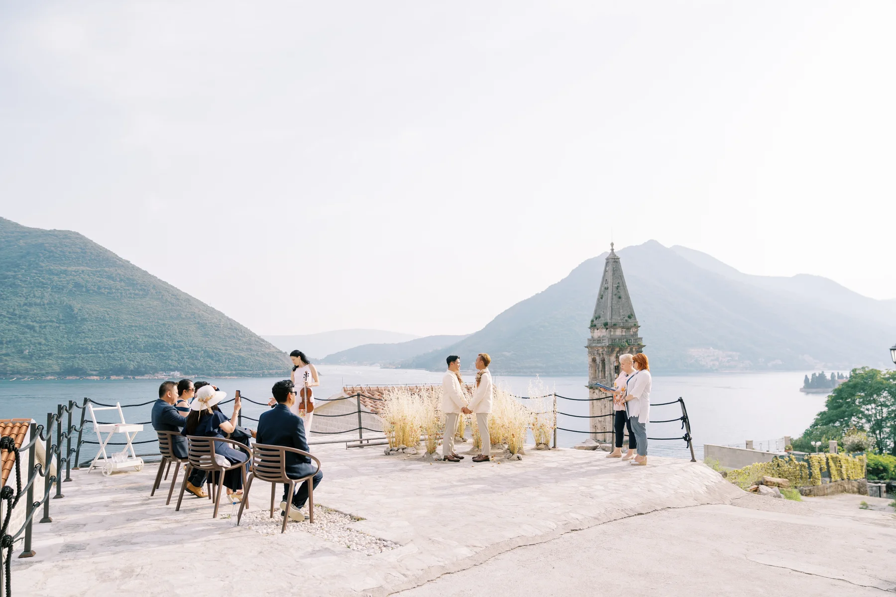 Ryan Jake and Jori exchange vows on a stone terrace at Monte Bay Retreat in Perast, Montenegro, with mountains and bay behind them.