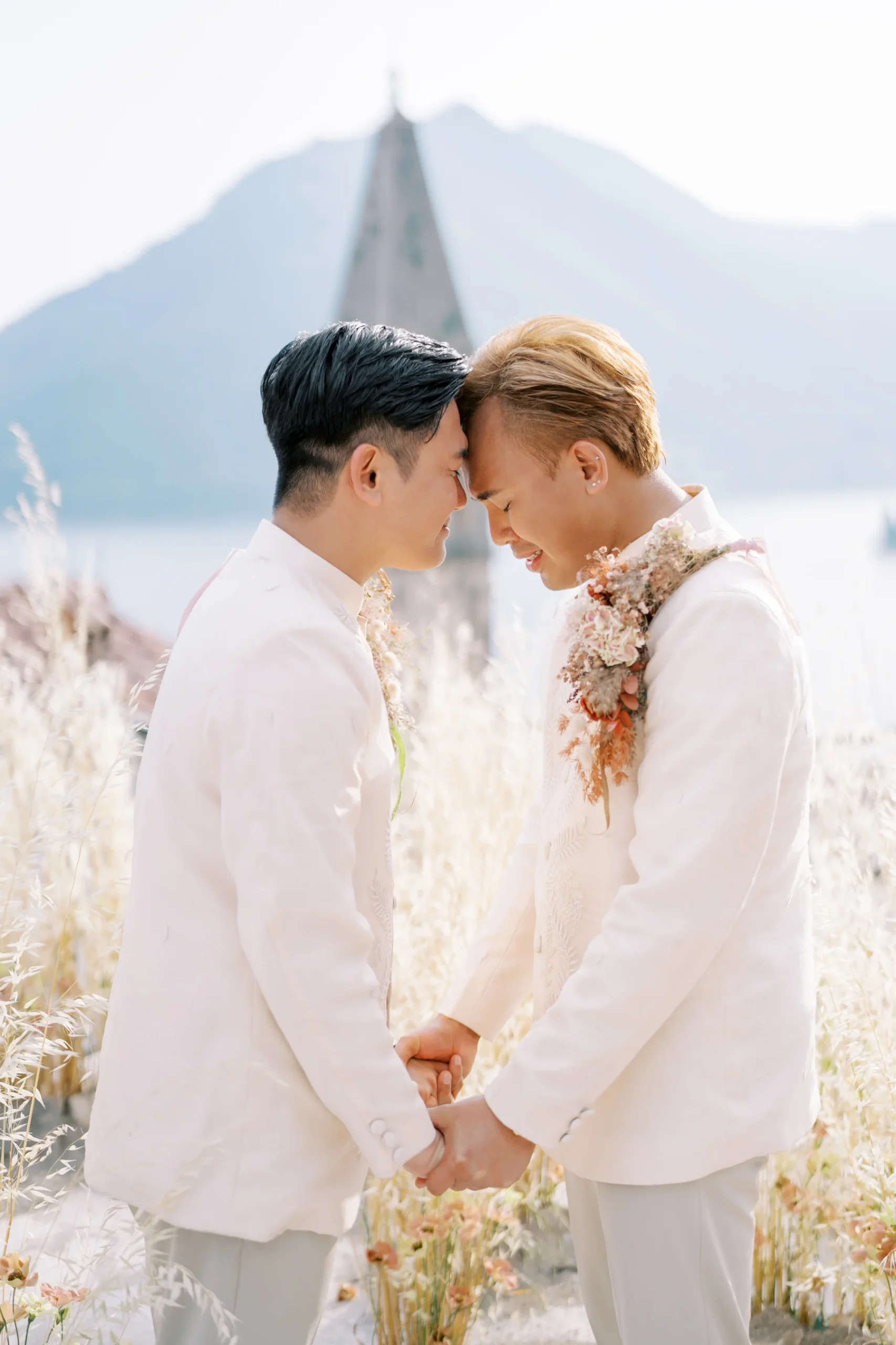 Ryan Jake and Jori hold hands, foreheads touching, in white suits amid pampas grass at Monte Bay Retreat in Perast, Montenegro.