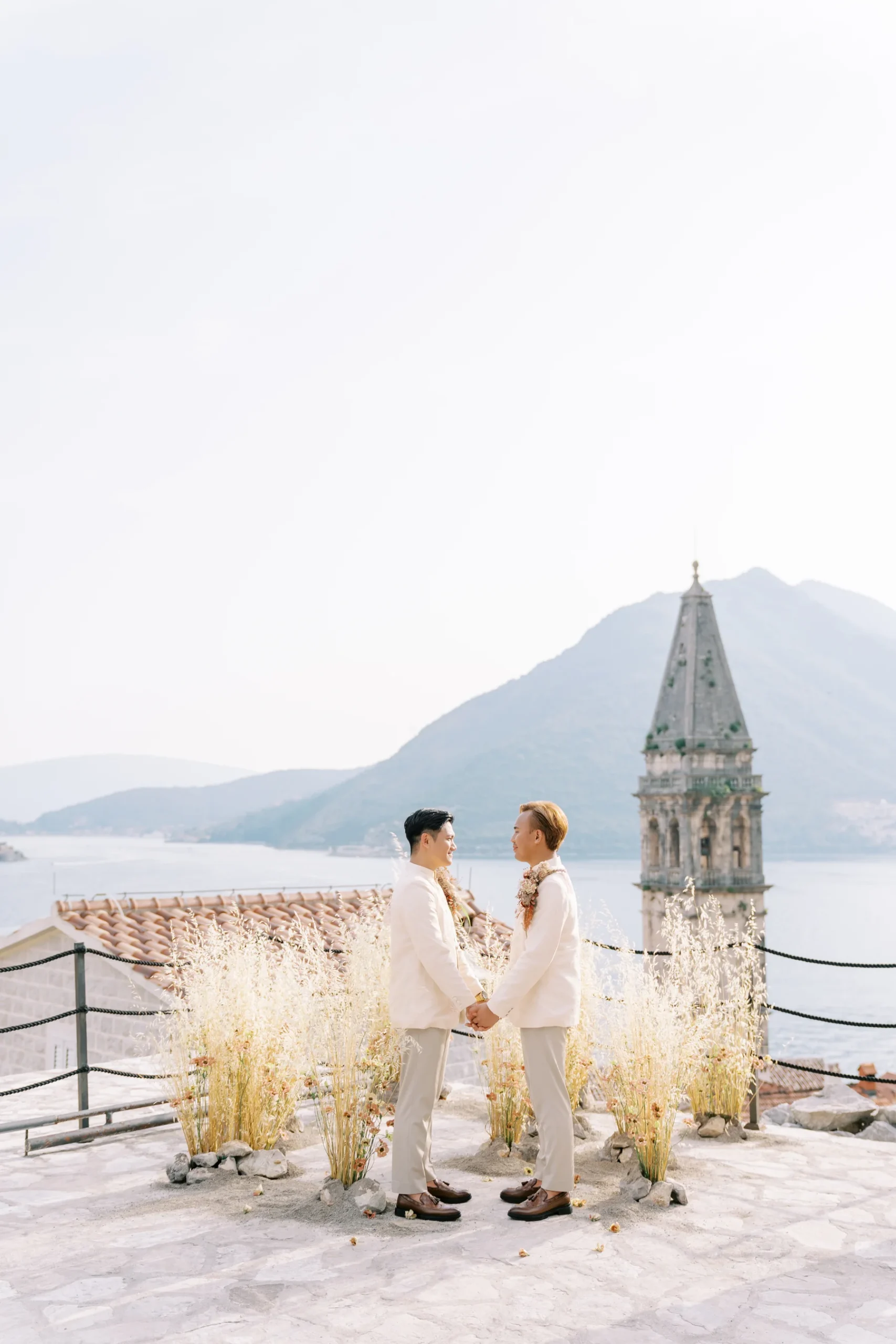 Ryan Jake and Jori hold hands at their outdoor ceremony at Monte Bay Retreat in Perast, Montenegro, with a church tower and bay behind them.