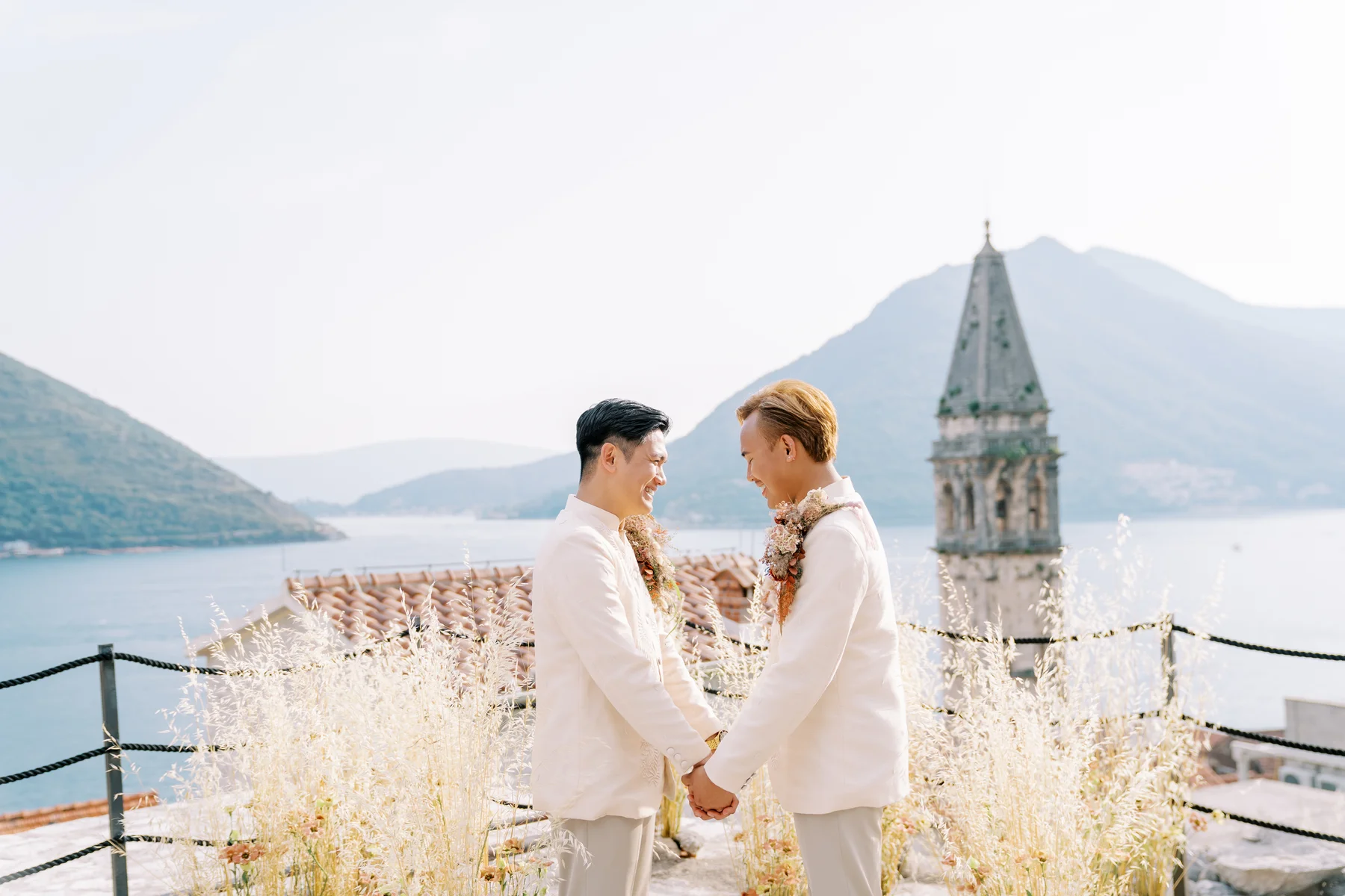 Ryan Jake and Jori hold hands in white suits with floral garlands at Monte Bay Retreat in Perast, Montenegro.