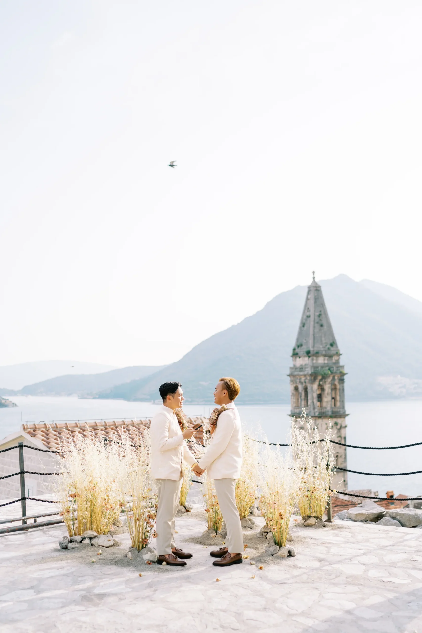 Ryan Jake and Jori exchange vows in white suits at Monte Bay Retreat in Perast, Montenegro, with a church tower and bay behind them.