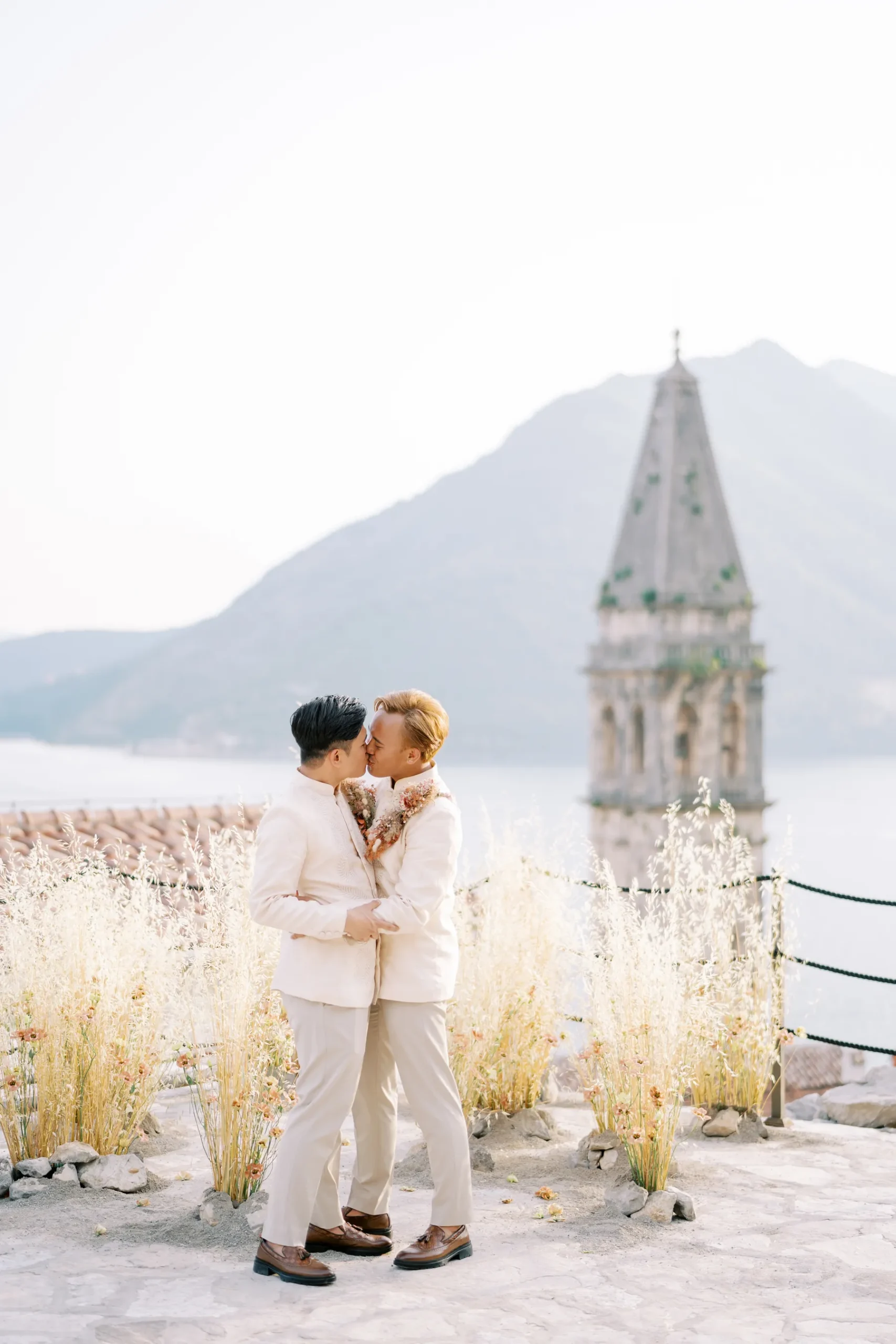Ryan Jake and Jori kiss in matching white suits at Our Lady of the Rocks island, Perast, Montenegro, with a stone bell tower behind them.