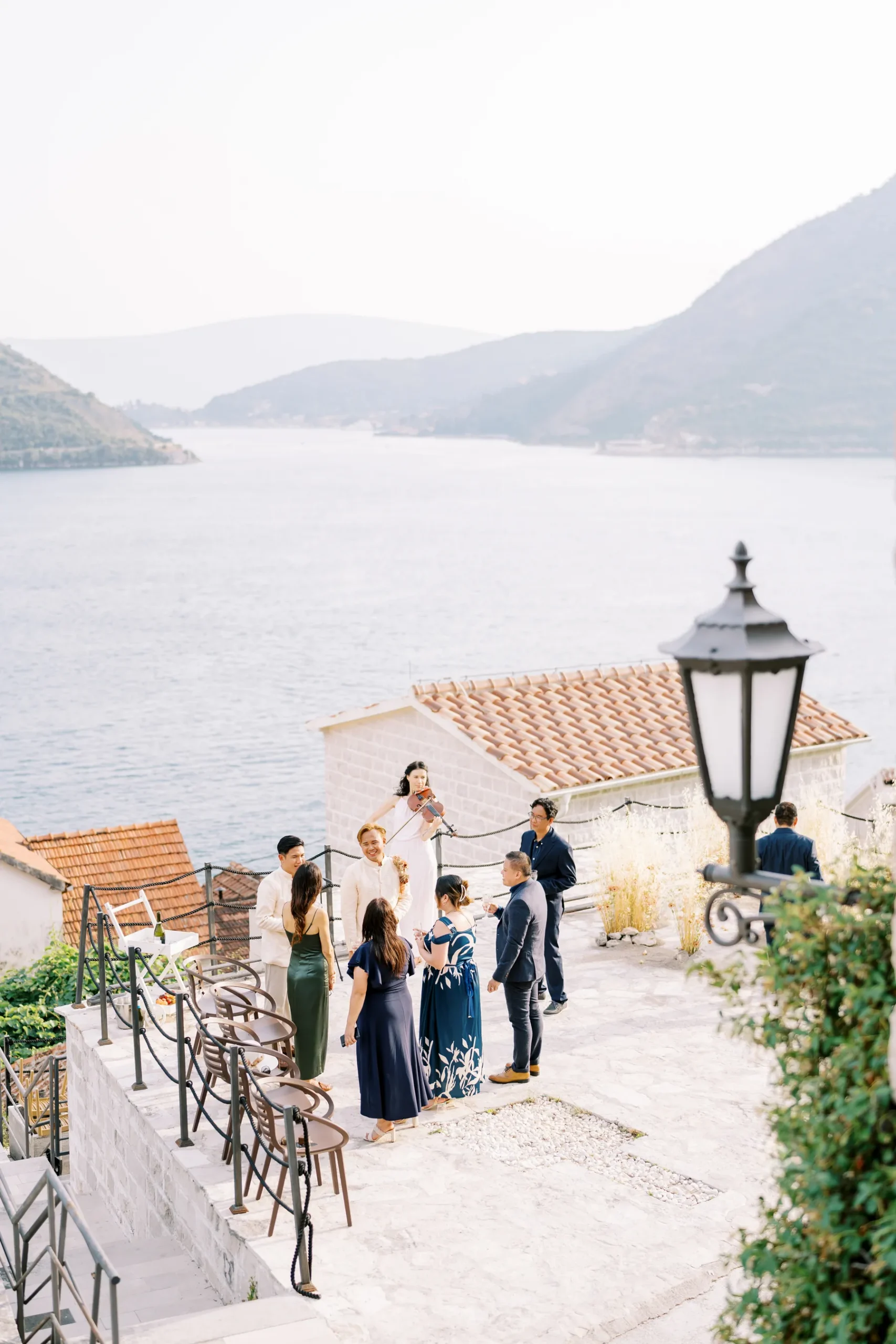 Wedding guests mingle on a stone terrace at Monte Bay Retreat in Perast, Montenegro, with a violinist performing and bay views behind them.