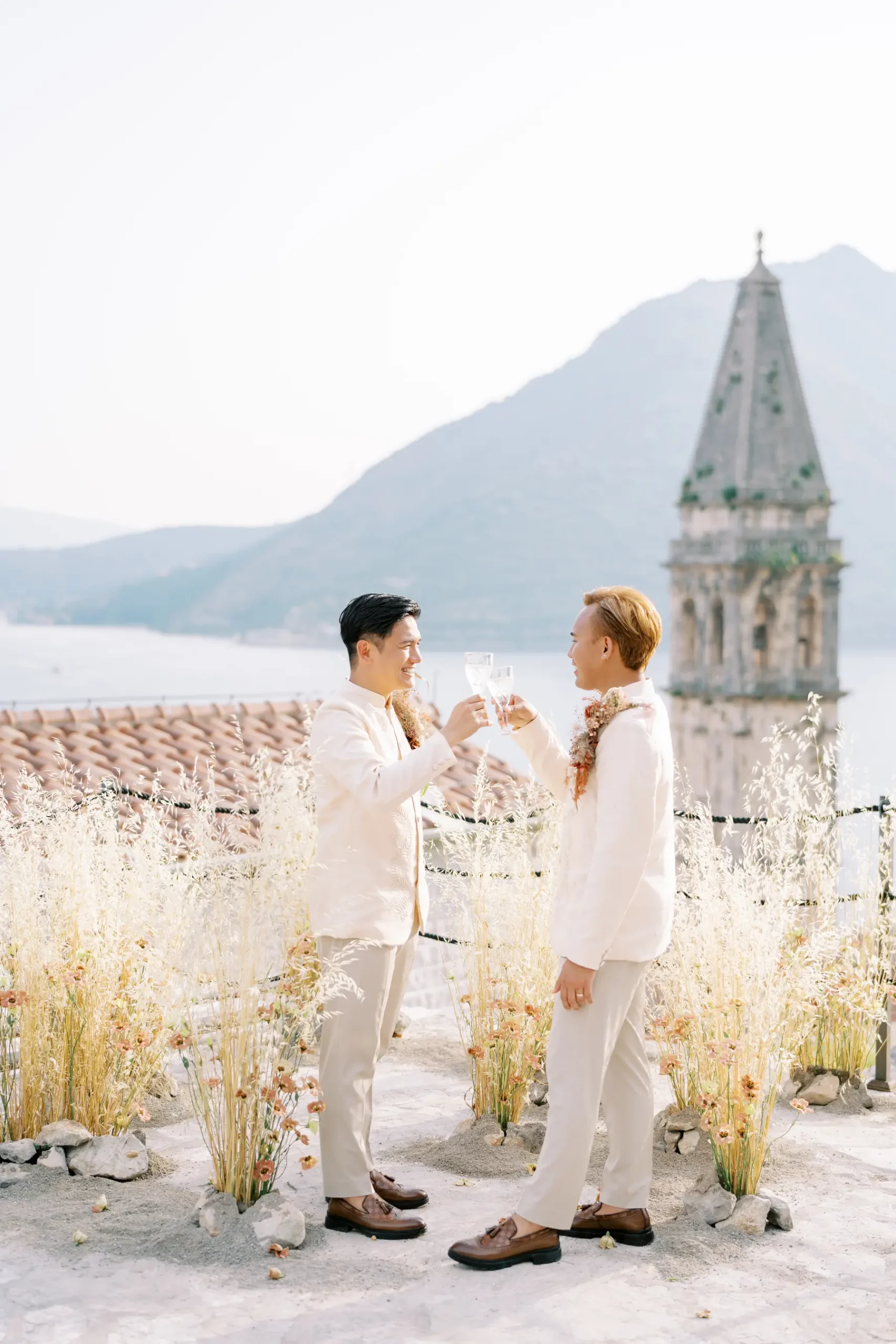 Ryan Jake and Jori toast with champagne in cream suits at Monte Bay Retreat in Perast, Montenegro, with a church tower behind them.