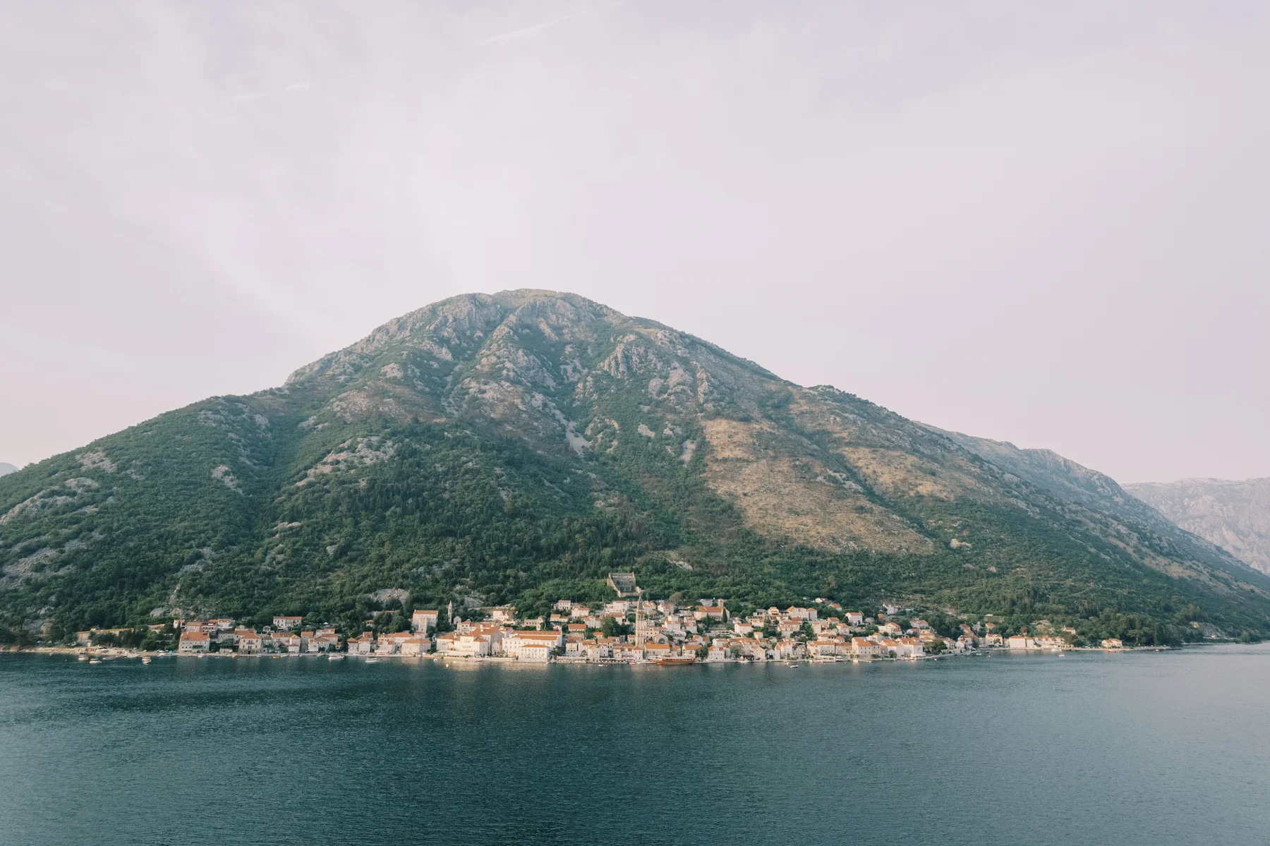 Panoramic view of Perast, Montenegro, from the Bay of Kotor, with terracotta-roofed buildings nestled at the base of a green mountain.
