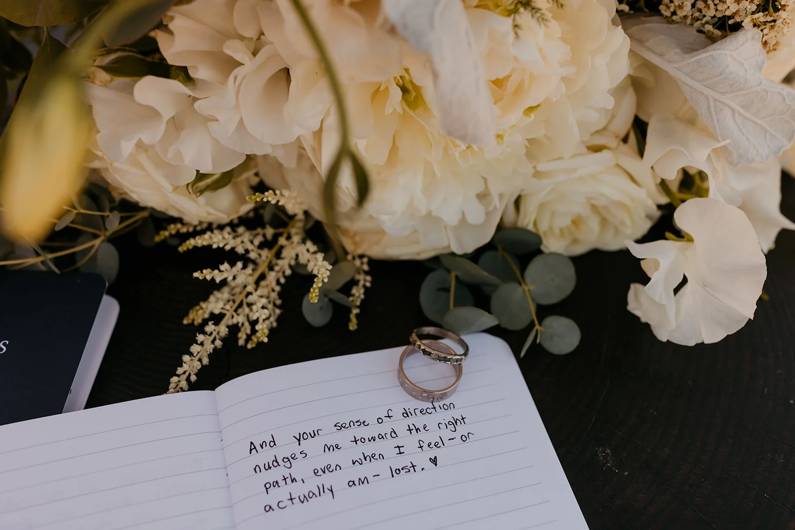 Wedding rings rest on a handwritten vows notebook beside white peonies and roses at Mayer State Park in Mosier, OR.