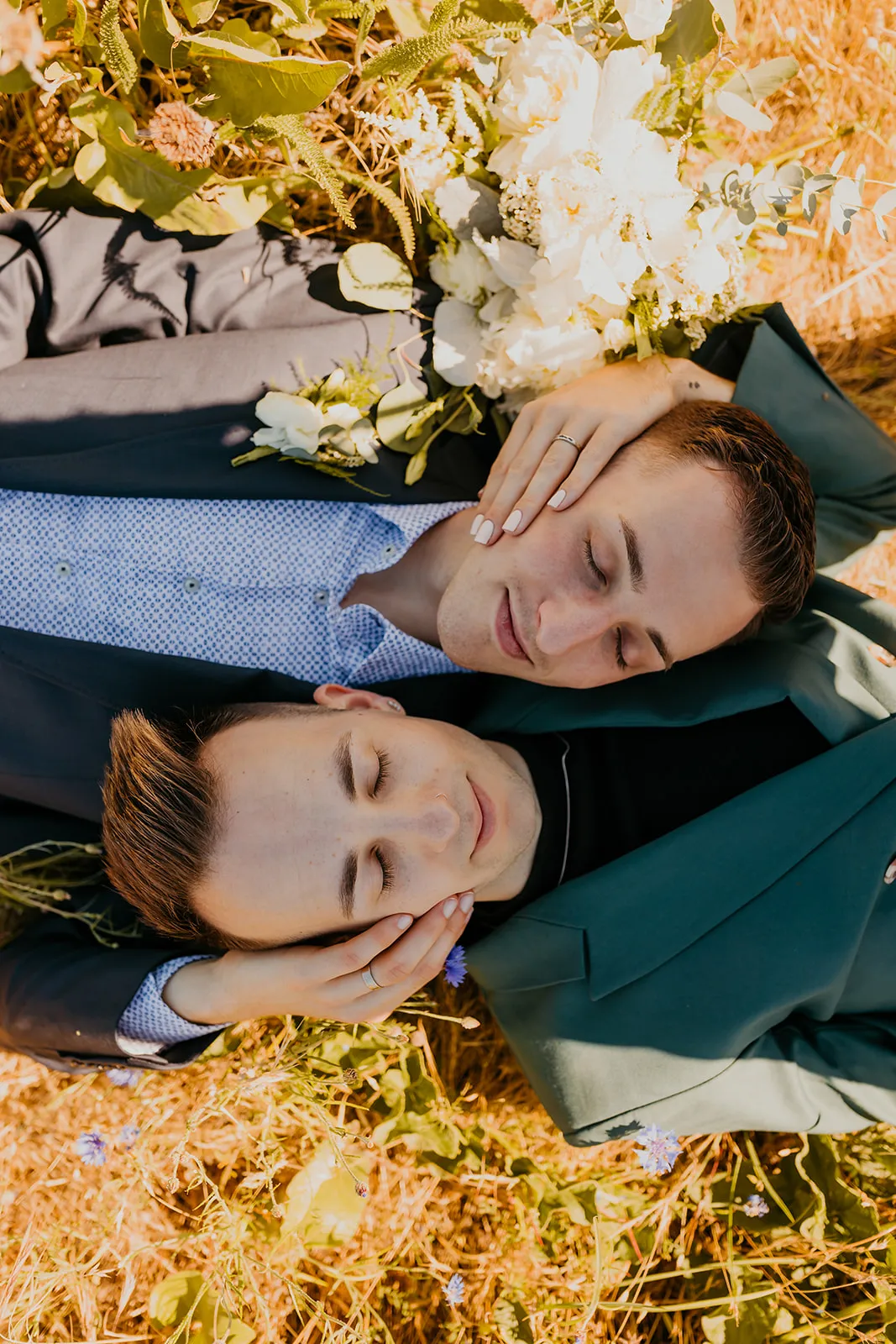 Ben and Nolan lie in golden grass at Mayer State Park, eyes closed, resting heads in each other's hands beside a bouquet of white roses, peonies, sweet peas, astilbe and greenery.
