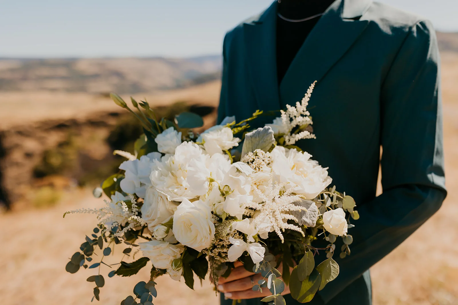 A lush white bridal bouquet of peonies and eucalyptus held by a person in a teal suit at Mayer State Park.