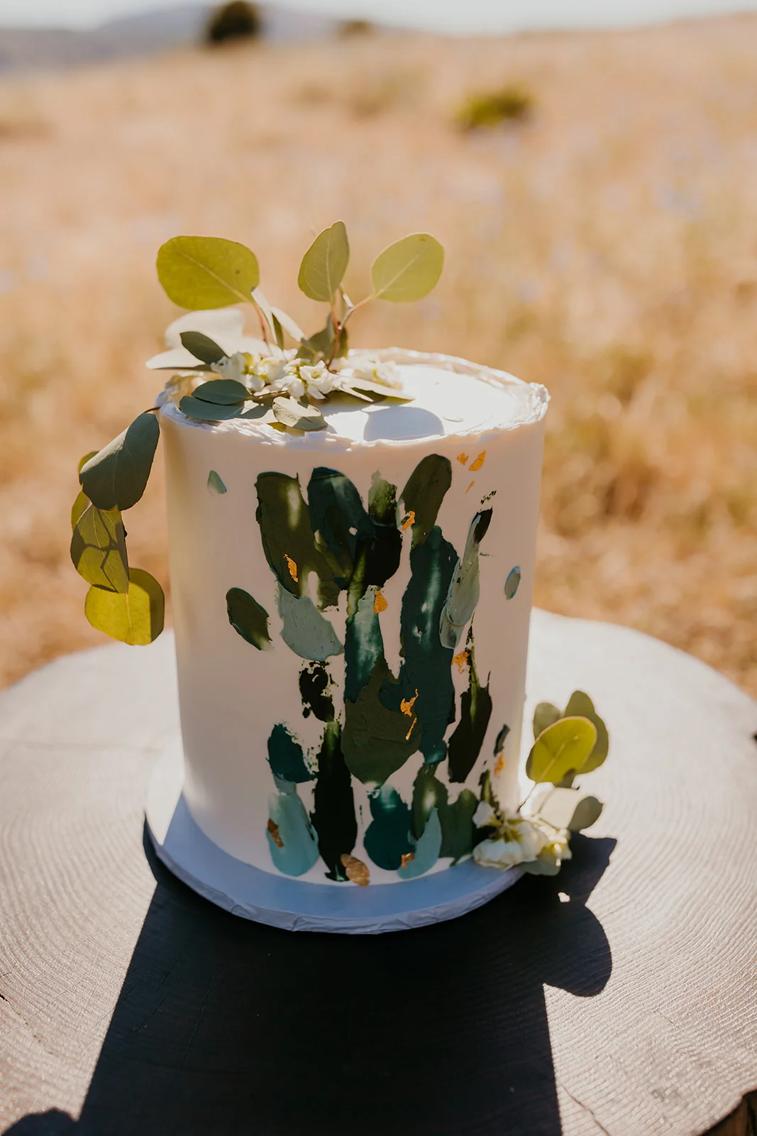 A white single-tier wedding cake with green brushstroke details and eucalyptus at Ben and Nolan's wedding at Mayer State Park.