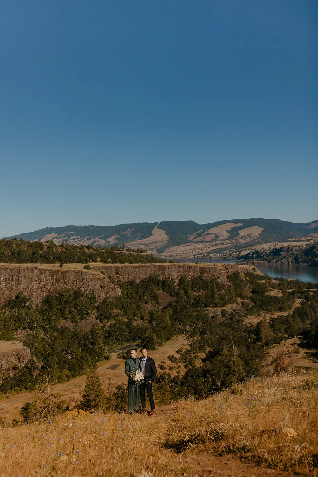Ben and Nolan stand together holding a bouquet on a hillside trail at Mayer State Park in Mosier, Oregon.