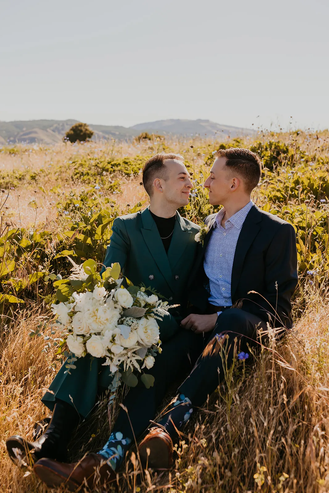 Ben and Nolan sit together in a sunlit wildflower field at Mayer State Park in Mosier, OR, gazing at each other.