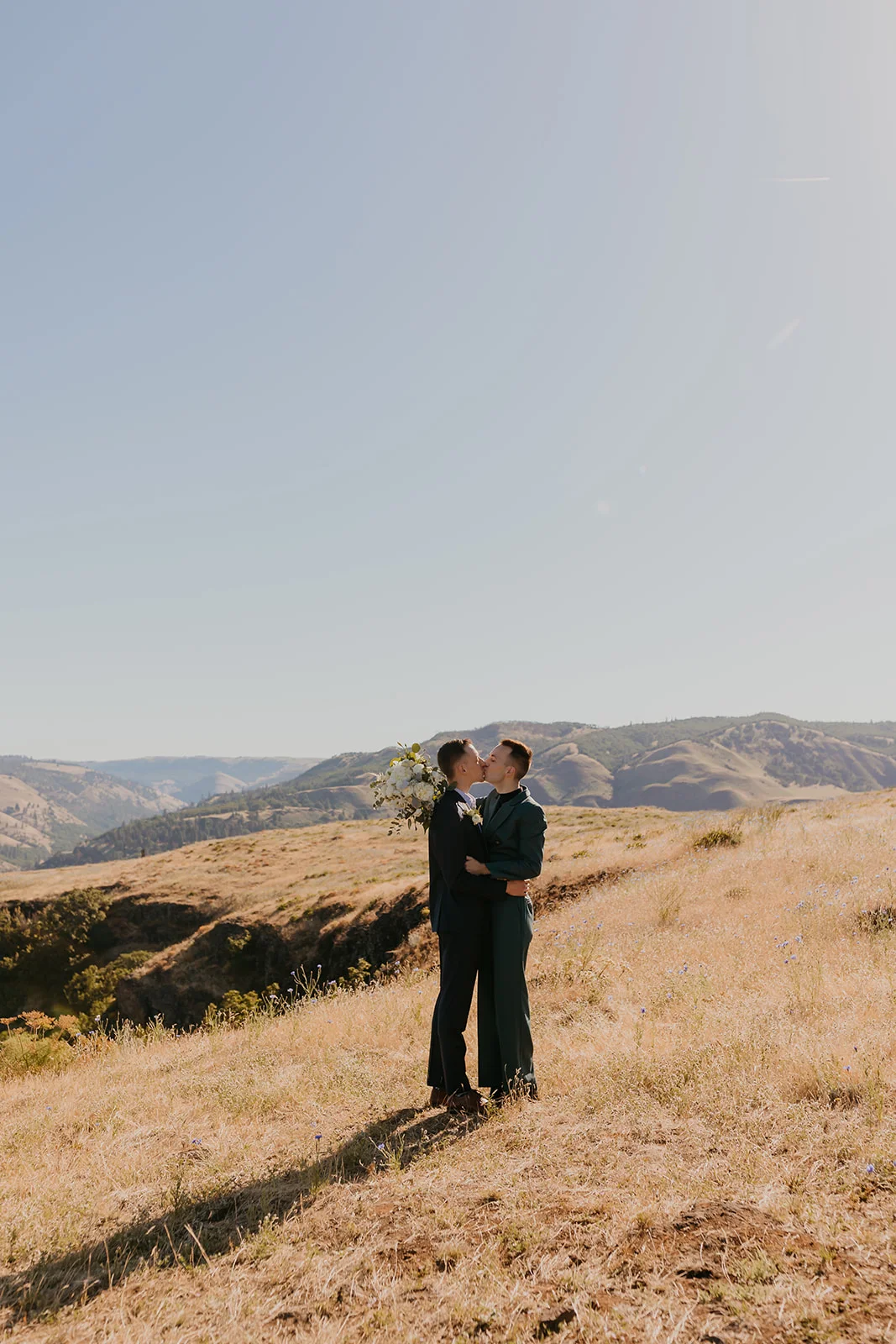 Ben and Nolan kiss on a golden hillside at Mayer State Park in Mosier, OR, holding a white floral bouquet.