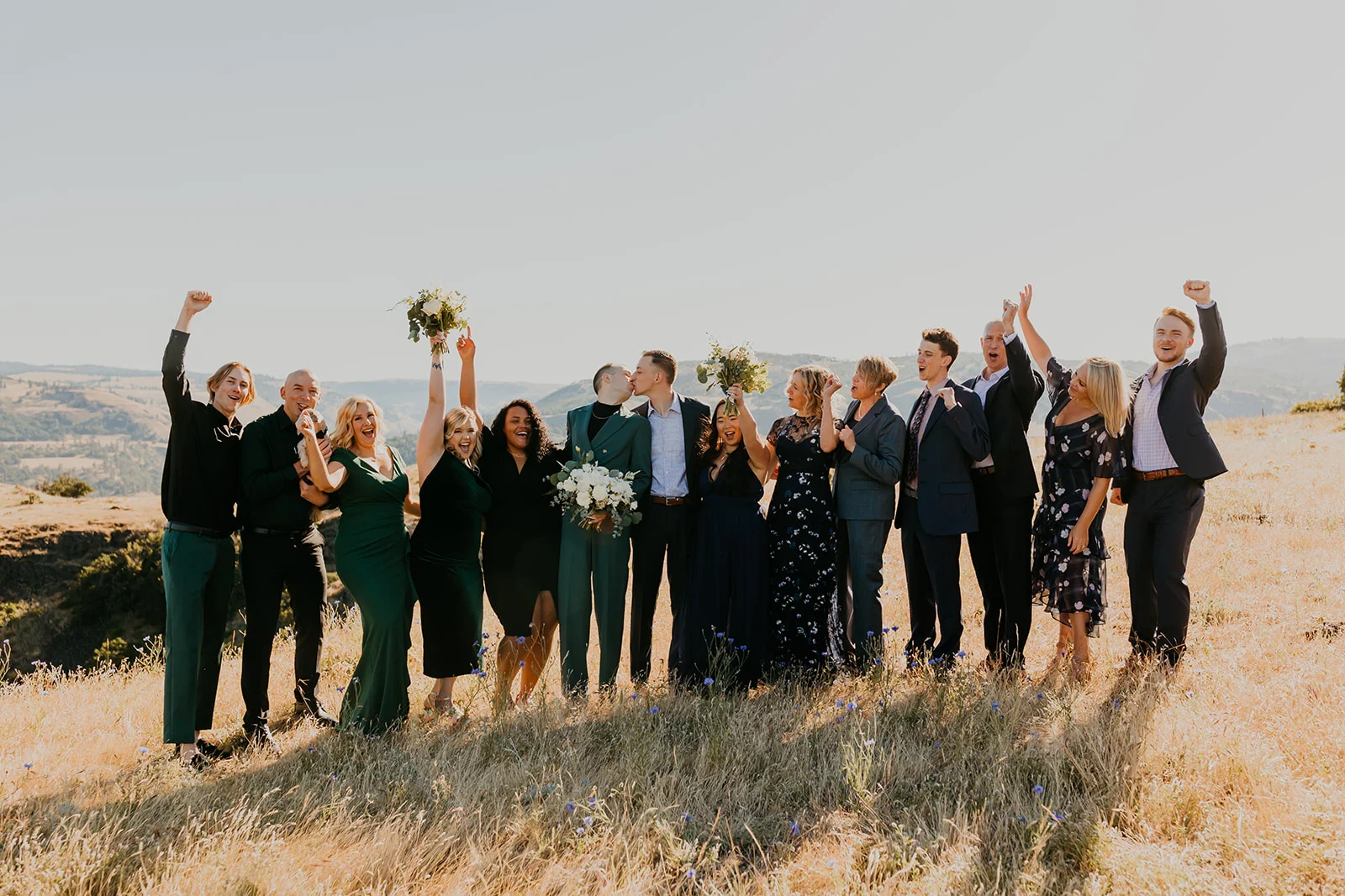 Ben and Nolan kiss surrounded by cheering wedding party on a golden hillside at Mayer State Park in Mosier, OR.