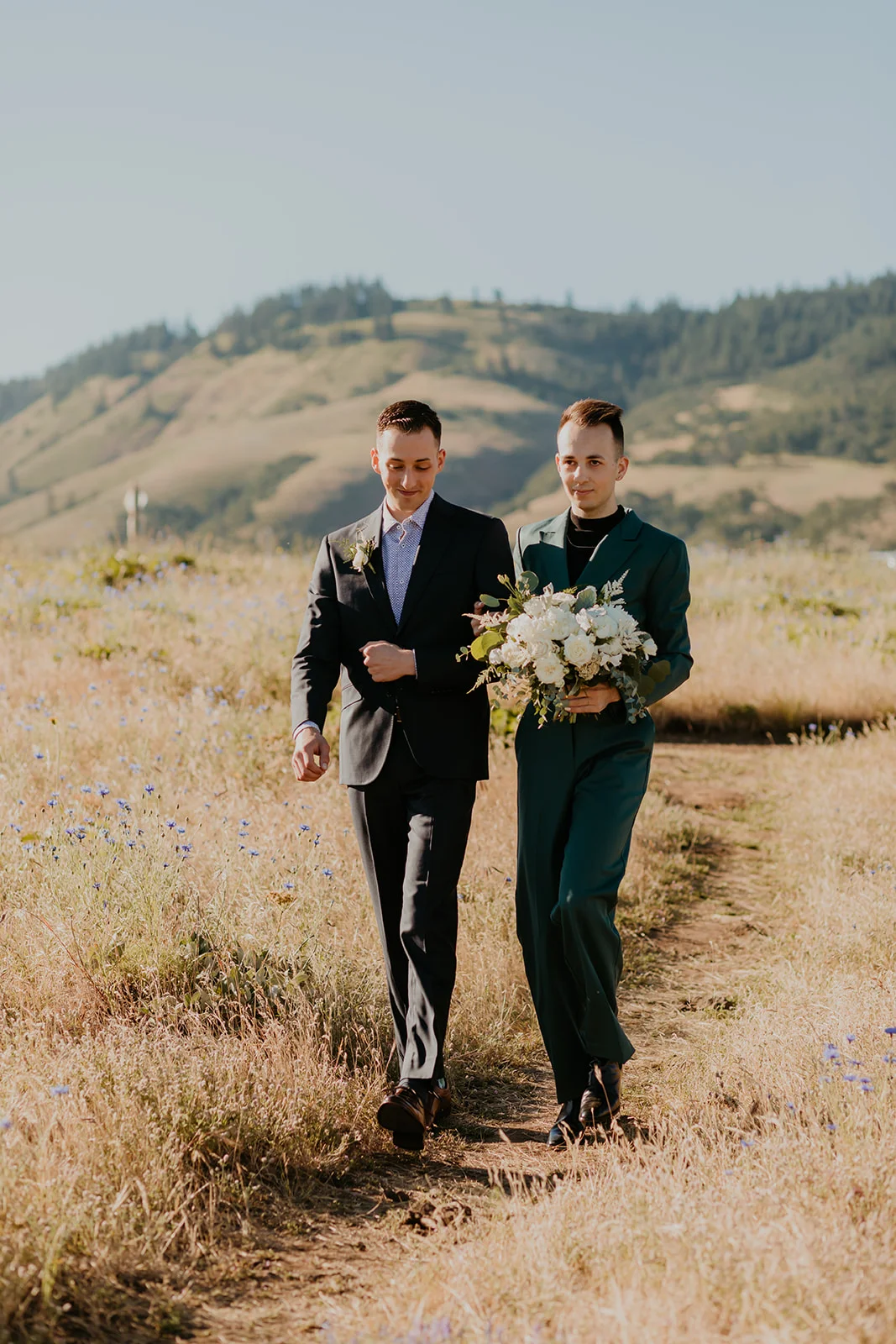 Ben and Nolan walk through a sunlit meadow at Mayer State Park in Mosier, OR, both in suits, one holding a white bouquet.