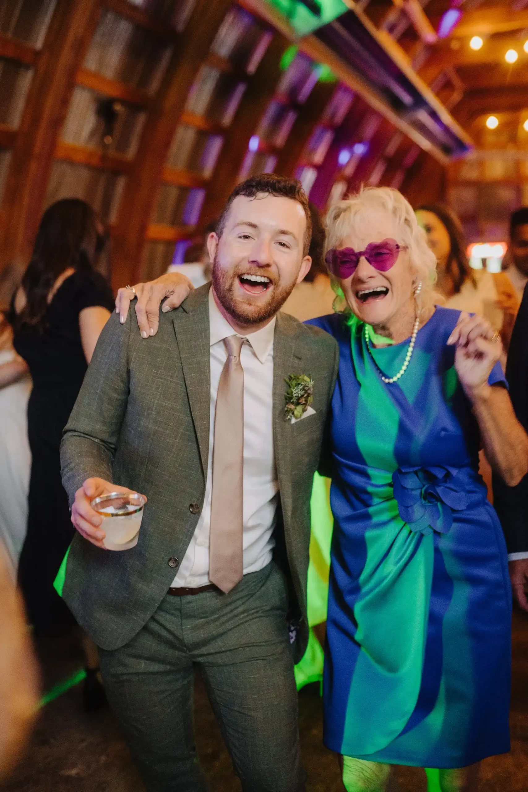 A laughing groom in a gray suit poses with an older woman in a blue-green dress and pink heart sunglasses at The Hillside Lodge.