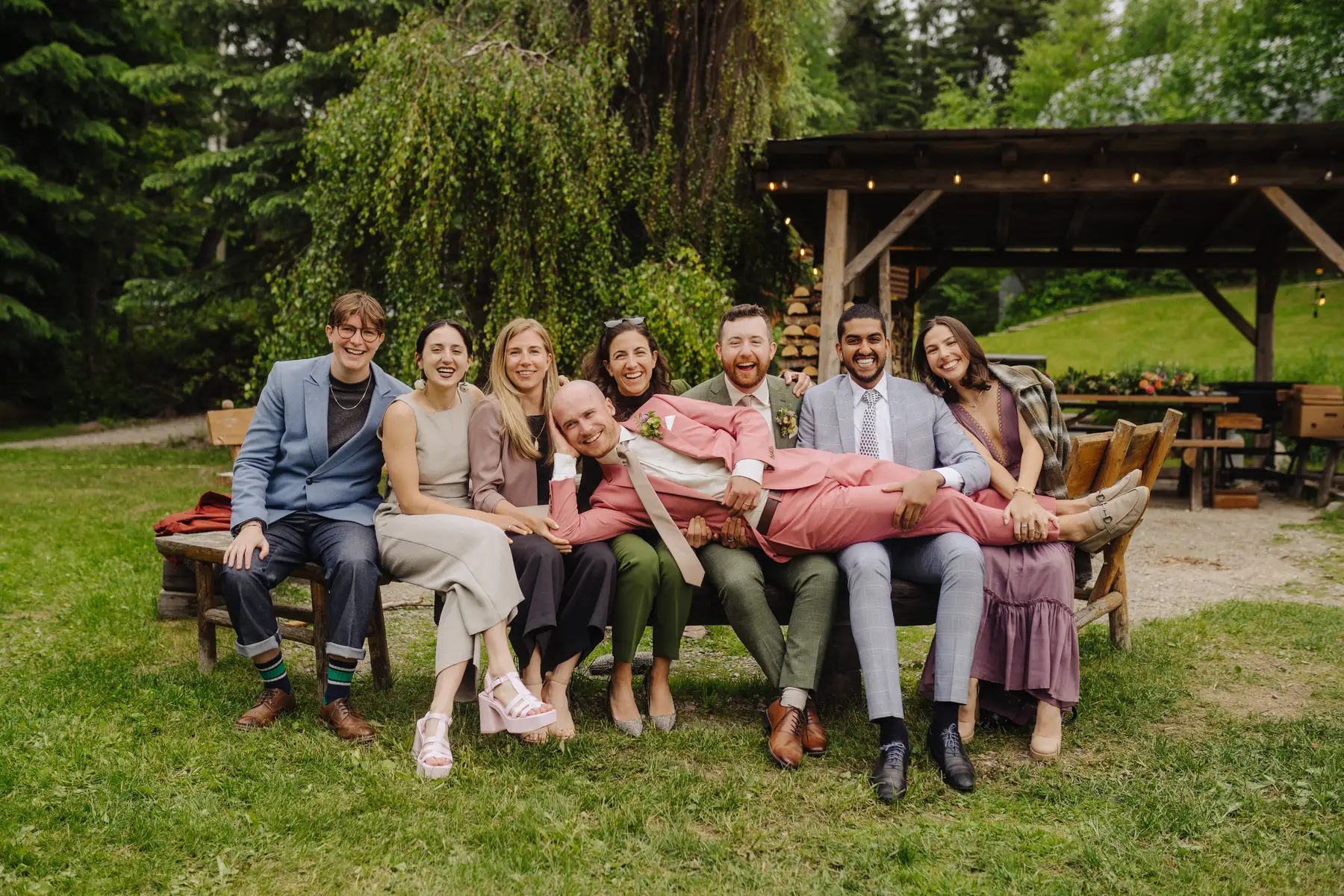 Wedding guests laugh as a man in a pink suit is held across their laps on a bench at The Hillside Lodge in Golden, BC.