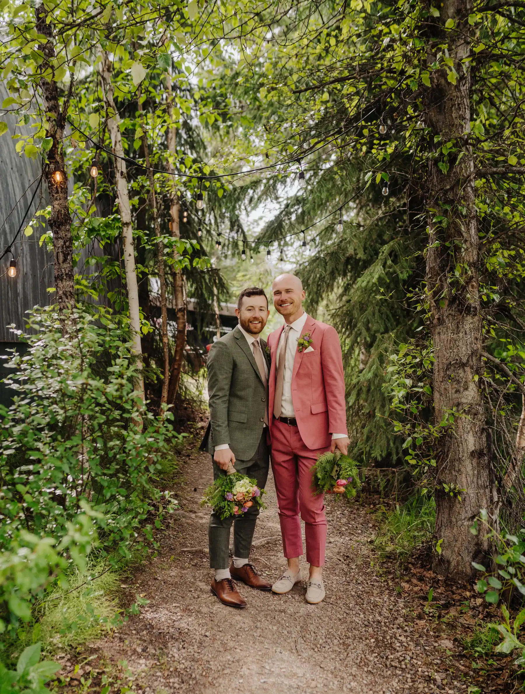 Trent and Cameron Harris-McCoy smile together on a wooded path at The Hillside Lodge & Chalets in Golden, BC, holding bouquets.