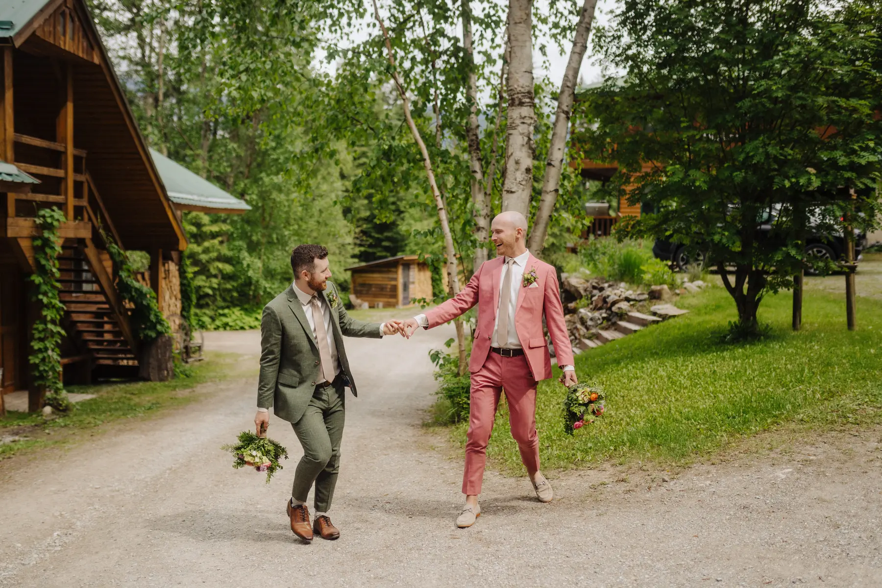 Trent and Cameron Harris-McCoy laugh and hold hands in coordinating suits outside The Hillside Lodge & Chalets in Golden, BC.