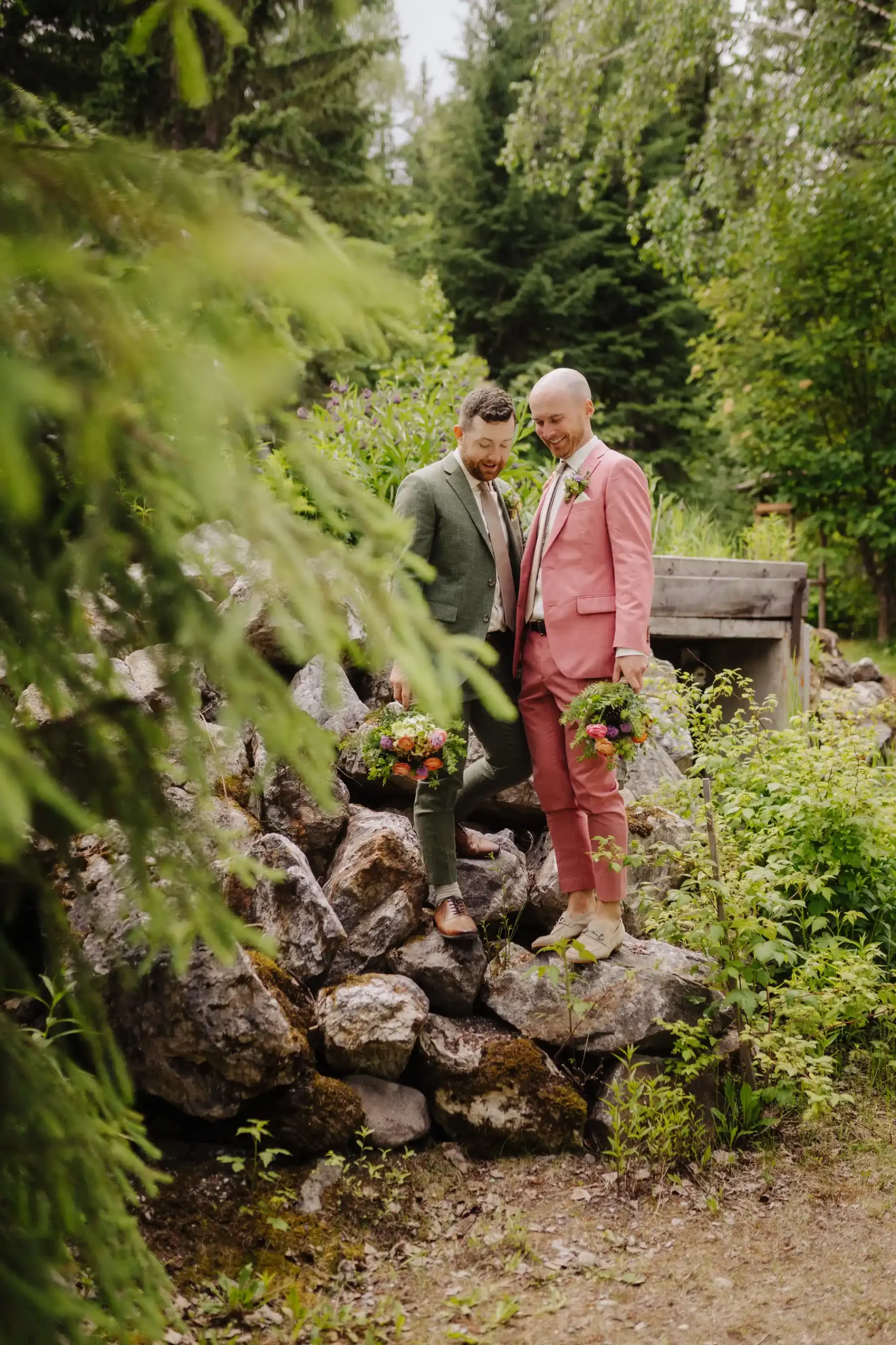 Trent and Cameron Harris-McCoy laugh together on mossy rocks holding bouquets at The Hillside Lodge in Golden, BC.