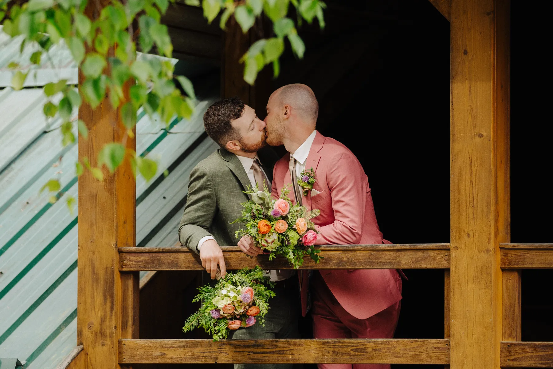 Trent and Cameron Harris-McCoy kiss on a wooden balcony at The Hillside Lodge & Chalets in Golden, BC, holding colorful bouquets.