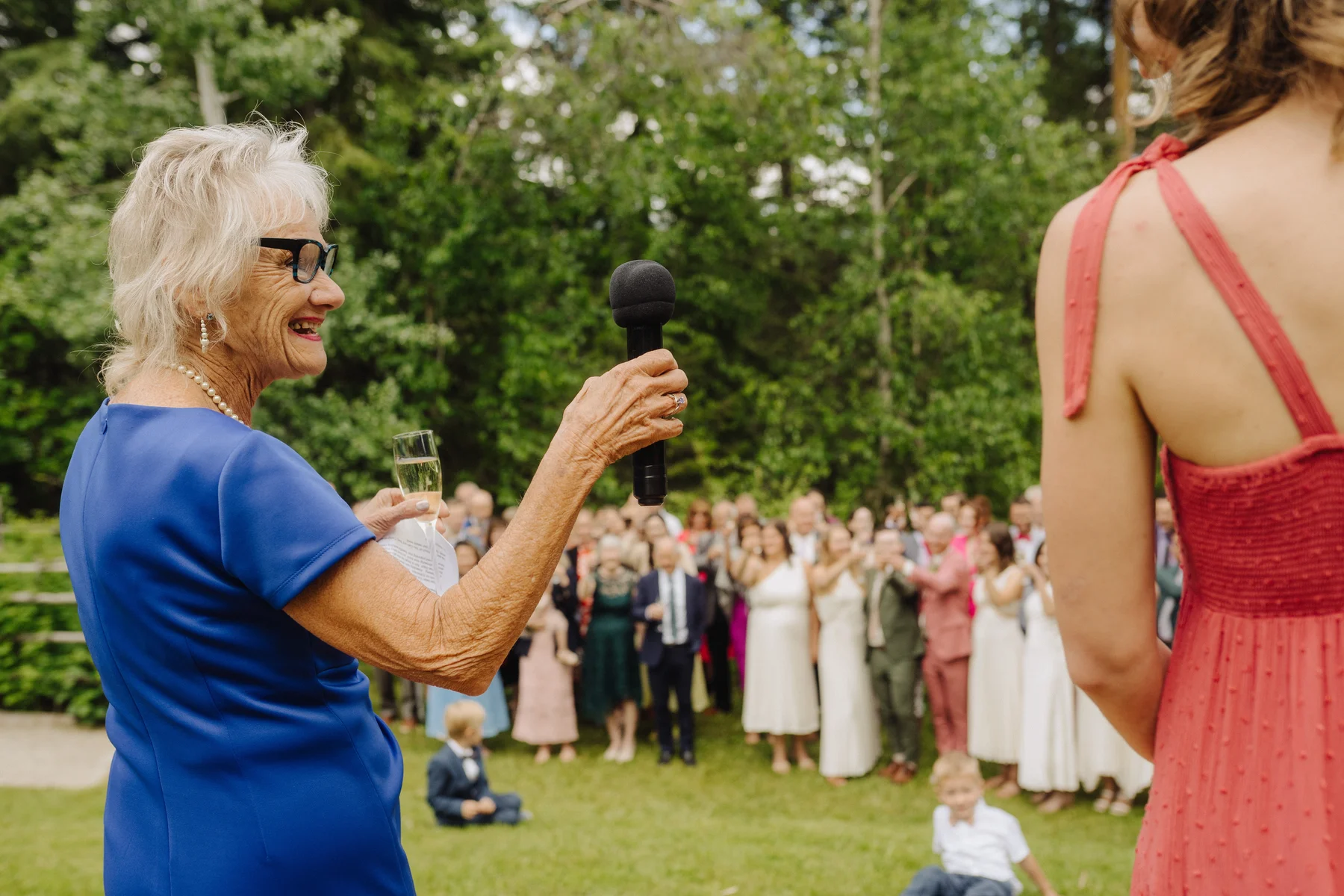 An older woman in a blue dress holds a microphone and champagne glass while giving a speech at an outdoor wedding reception.