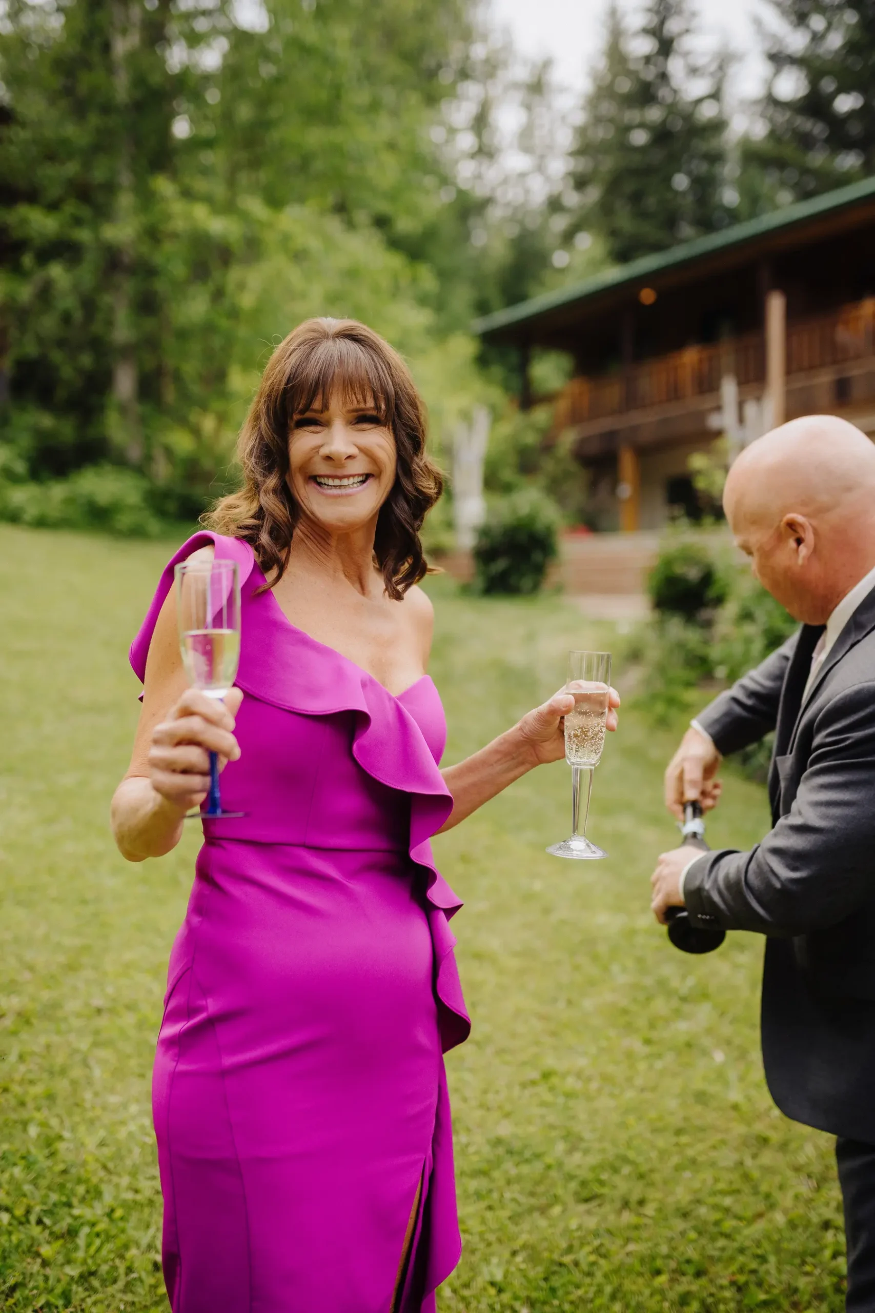 A smiling wedding guest in a magenta dress holds two champagne flutes at The Hillside Lodge & Chalets in Golden, BC.