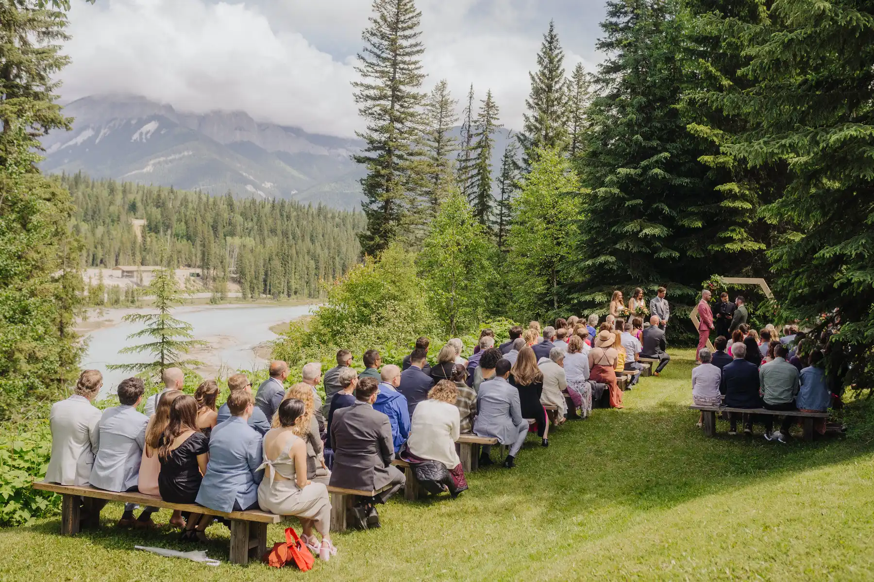 Guests seated on wooden benches watch Trent and Cameron Harris-McCoy's outdoor ceremony at The Hillside Lodge in Golden, BC, Canada.