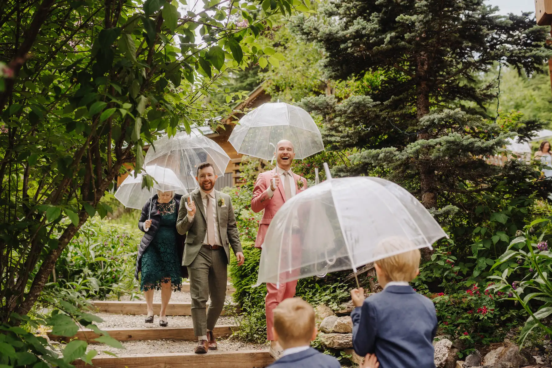 Trent and Cameron Harris-McCoy laugh walking down a garden path with clear umbrellas at The Hillside Lodge in Golden, BC.