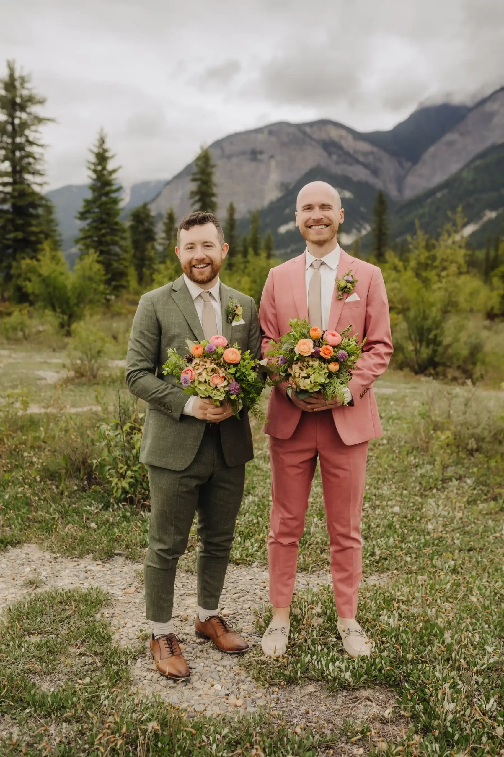 Trent and Cameron Harris-McCoy smile holding floral bouquets in suits at The Hillside Lodge & Chalets in Golden, BC.