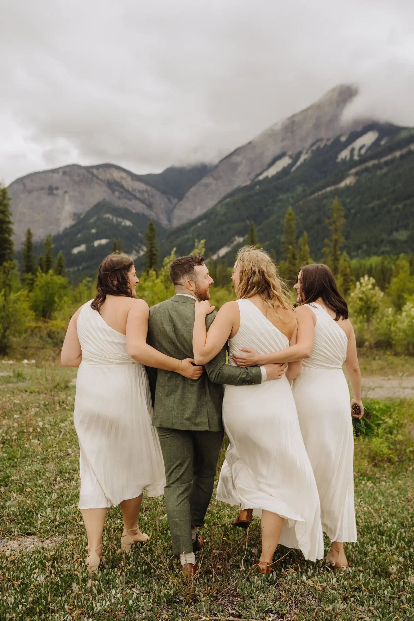 Trent and Cameron Harris-McCoy walk arm-in-arm with two bridesmaids in white dresses against a mountain backdrop in Golden, BC.