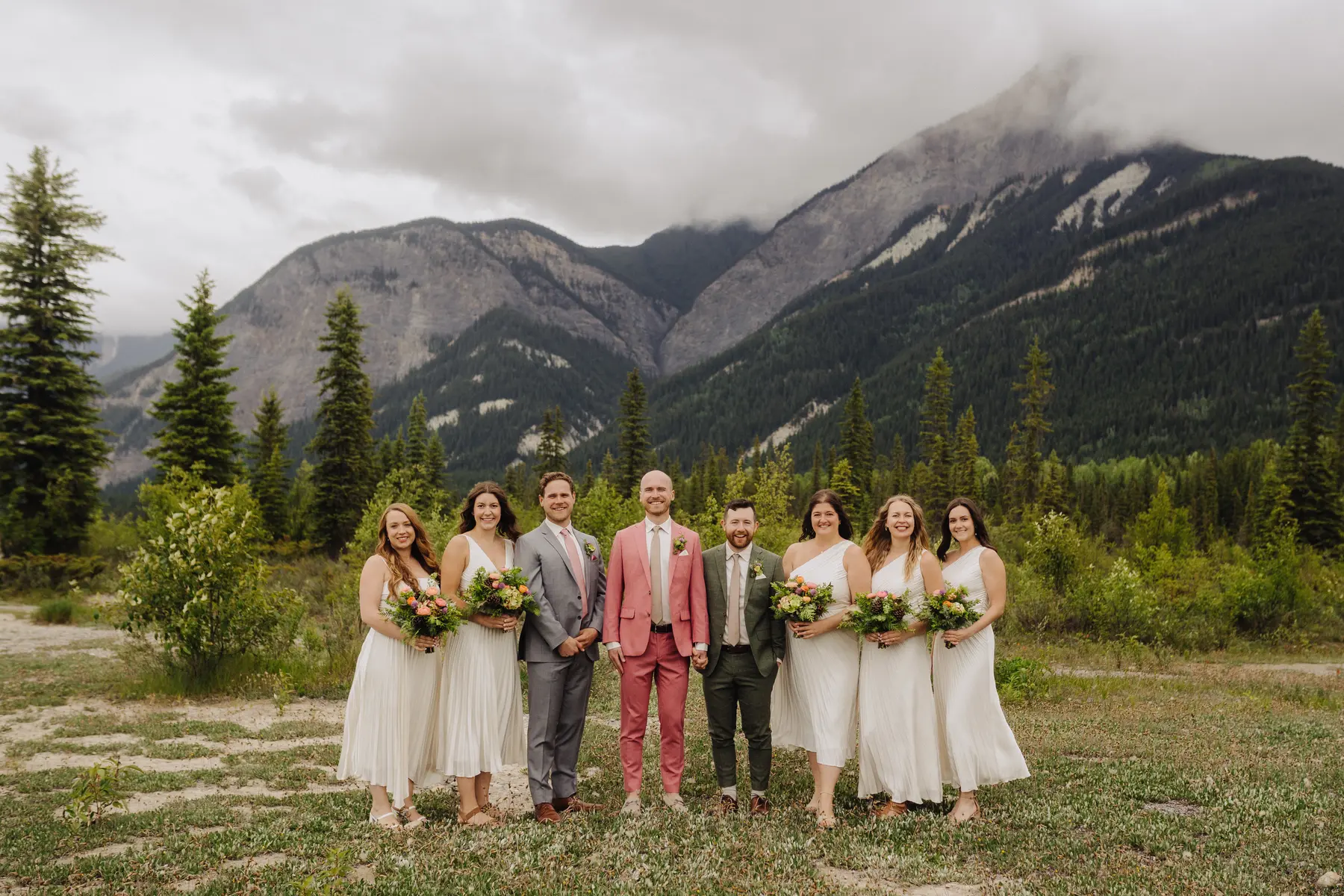 Trent and Cameron Harris-McCoy stand with their wedding party in white dresses and suits before misty mountains at The Hillside Lodge, Golden, BC.