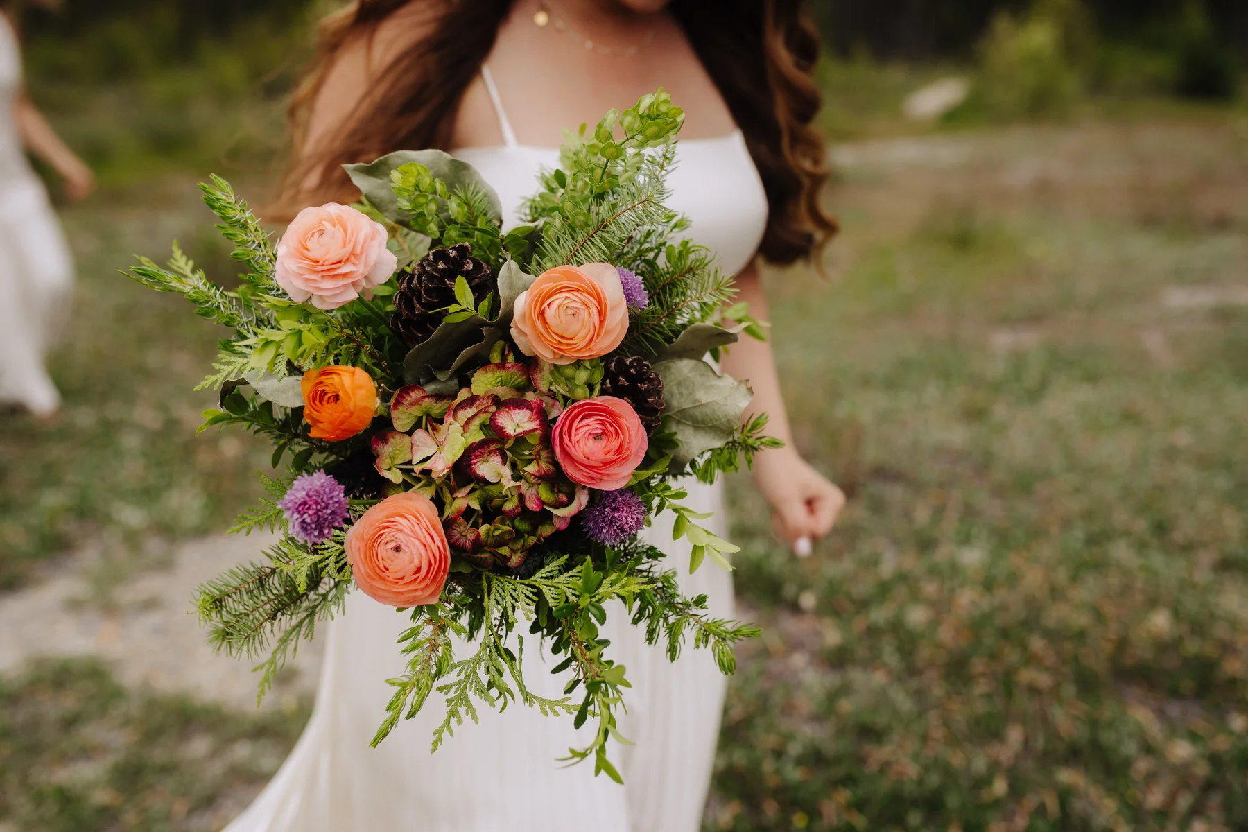 A bride in a white gown holds a lush bouquet of peach ranunculus, roses, hydrangeas and pine cones at The Hillside Lodge & Chalets.