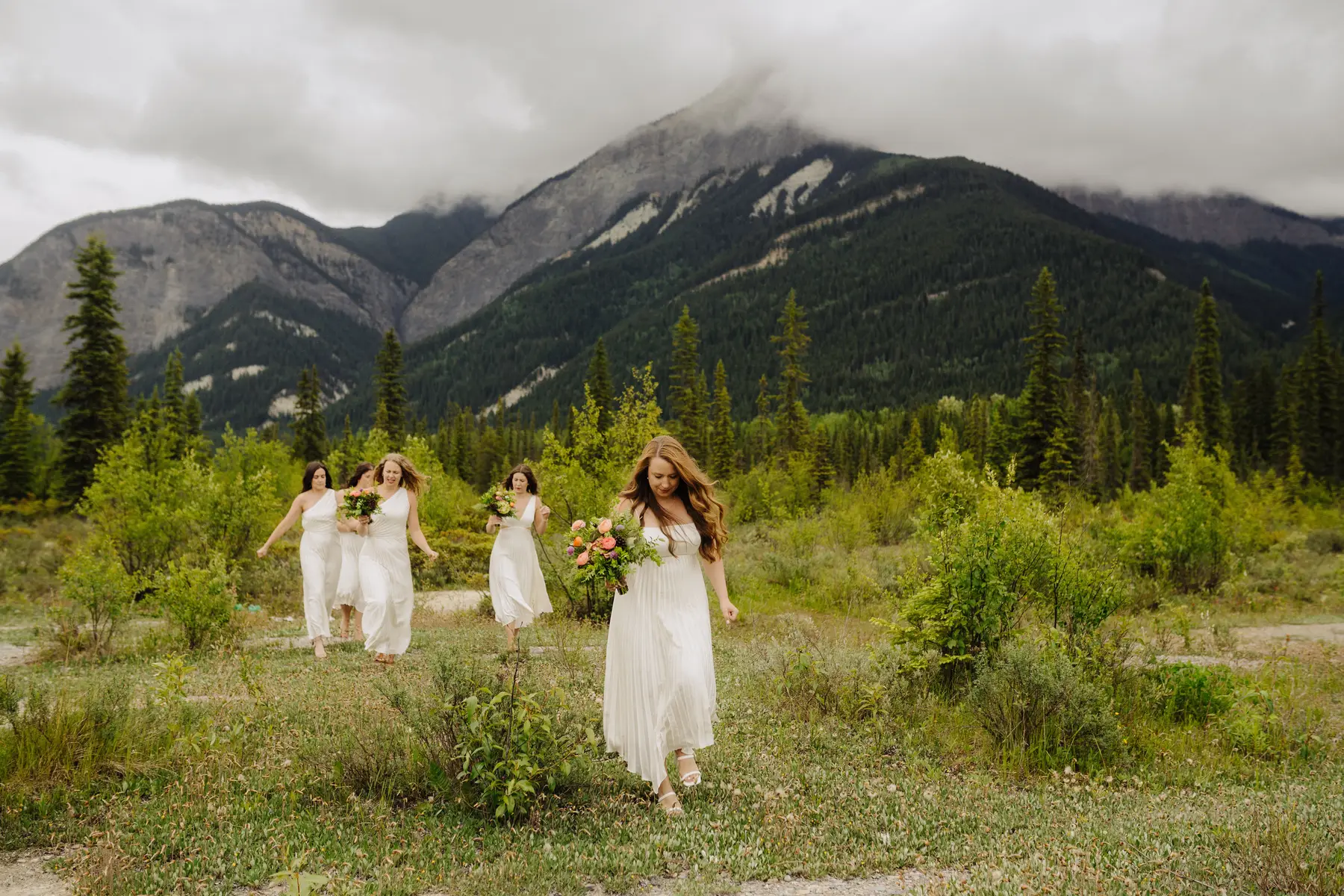 Bride leads bridesmaids in white dresses through a mountain meadow at The Hillside Lodge & Chalets in Golden, BC, Canada.