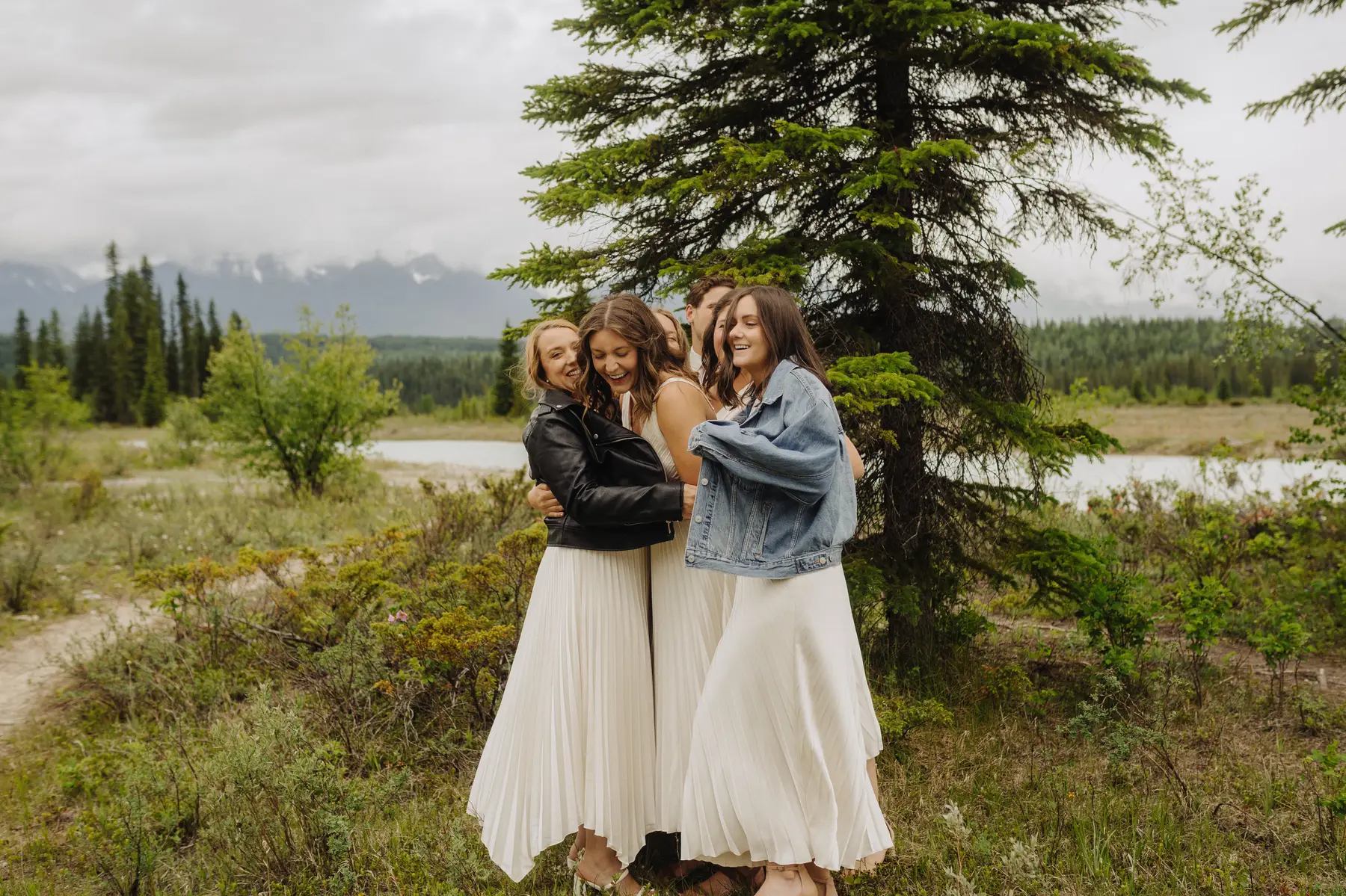 Bridesmaids in white pleated skirts laugh and embrace outdoors at The Hillside Lodge & Chalets in Golden, BC, Canada.