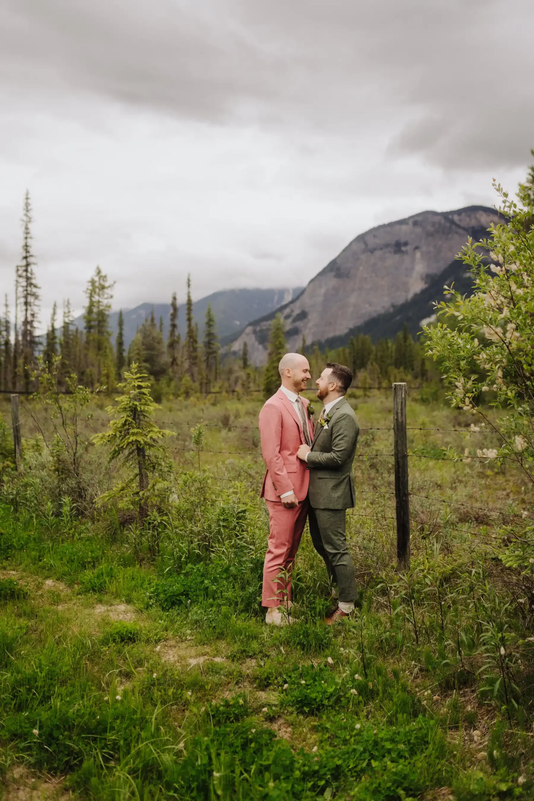Trent and Cameron Harris-McCoy share a joyful moment in suits beside a fence with mountain views at The Hillside Lodge, Golden, BC.