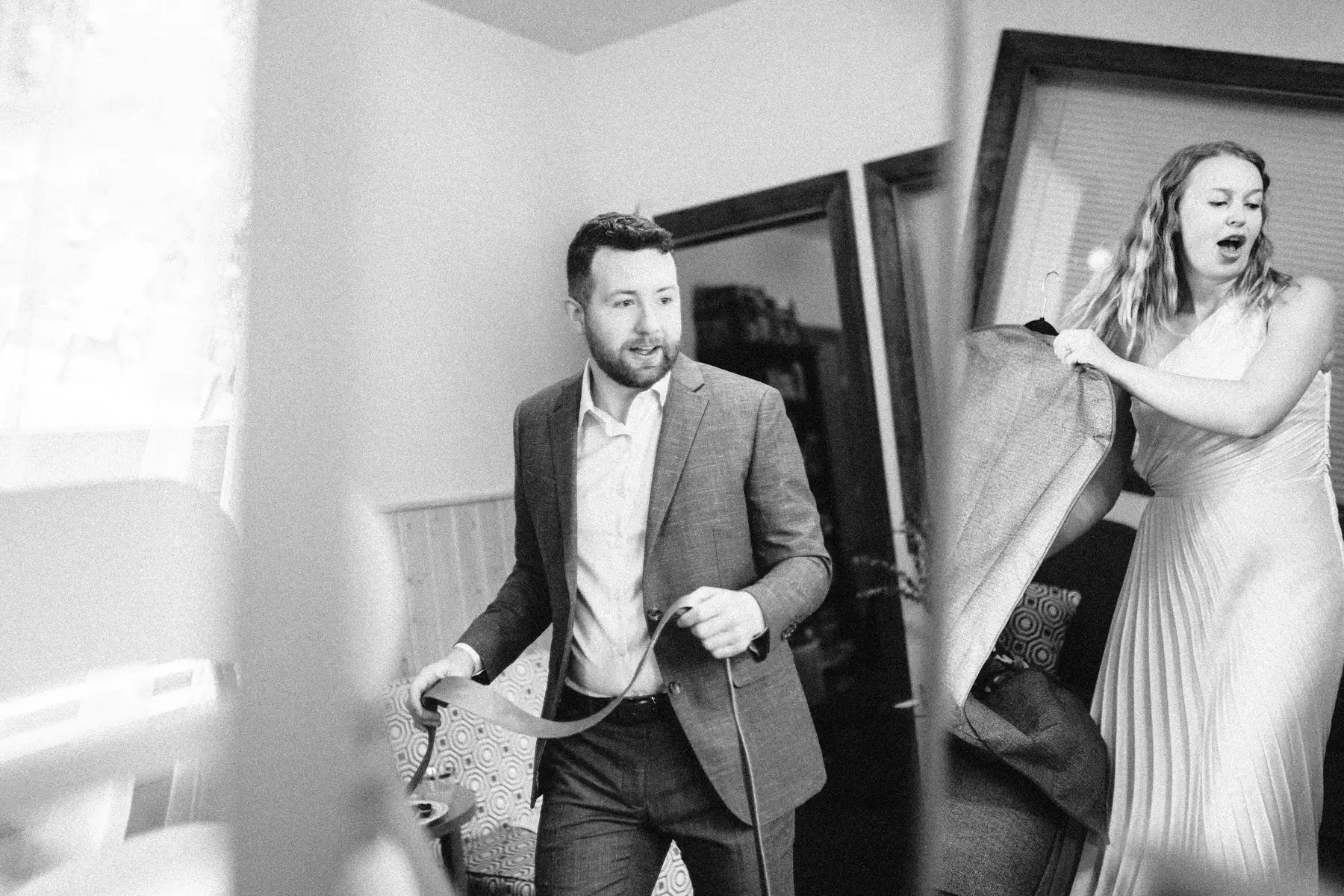 A man in a gray suit holds a tie while a woman in a flowy dress carries a garment bag inside The Hillside Lodge & Chalets.