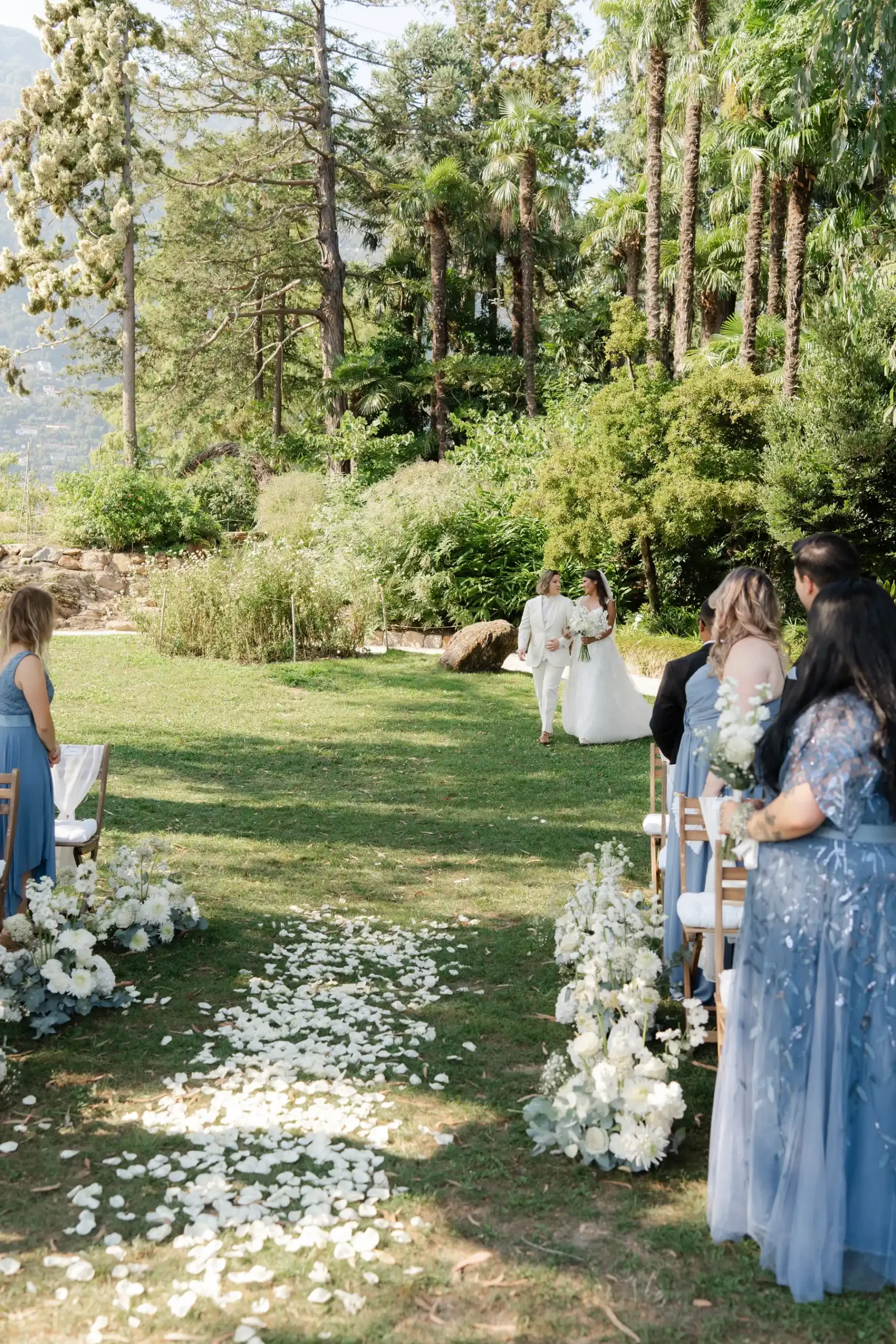 Ashley and Dionne walk down a flower-petal aisle at Villa Emden on Brissago Island, Switzerland, as guests in blue look on.