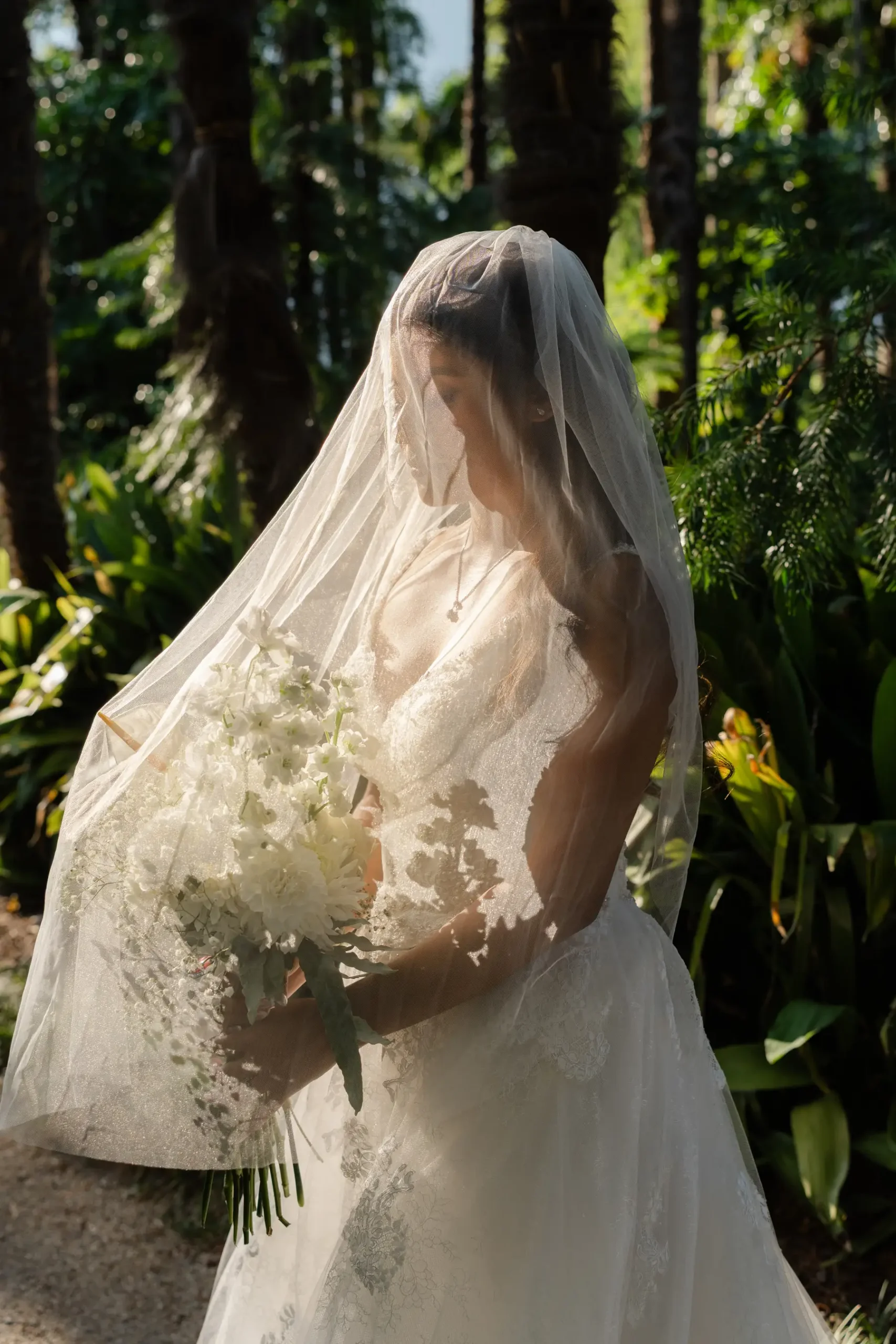 Bride Ashley Rodriguez-Vargas in a lace gown and veil holds a white bouquet amid lush gardens at Villa Emden, Brissago Island.