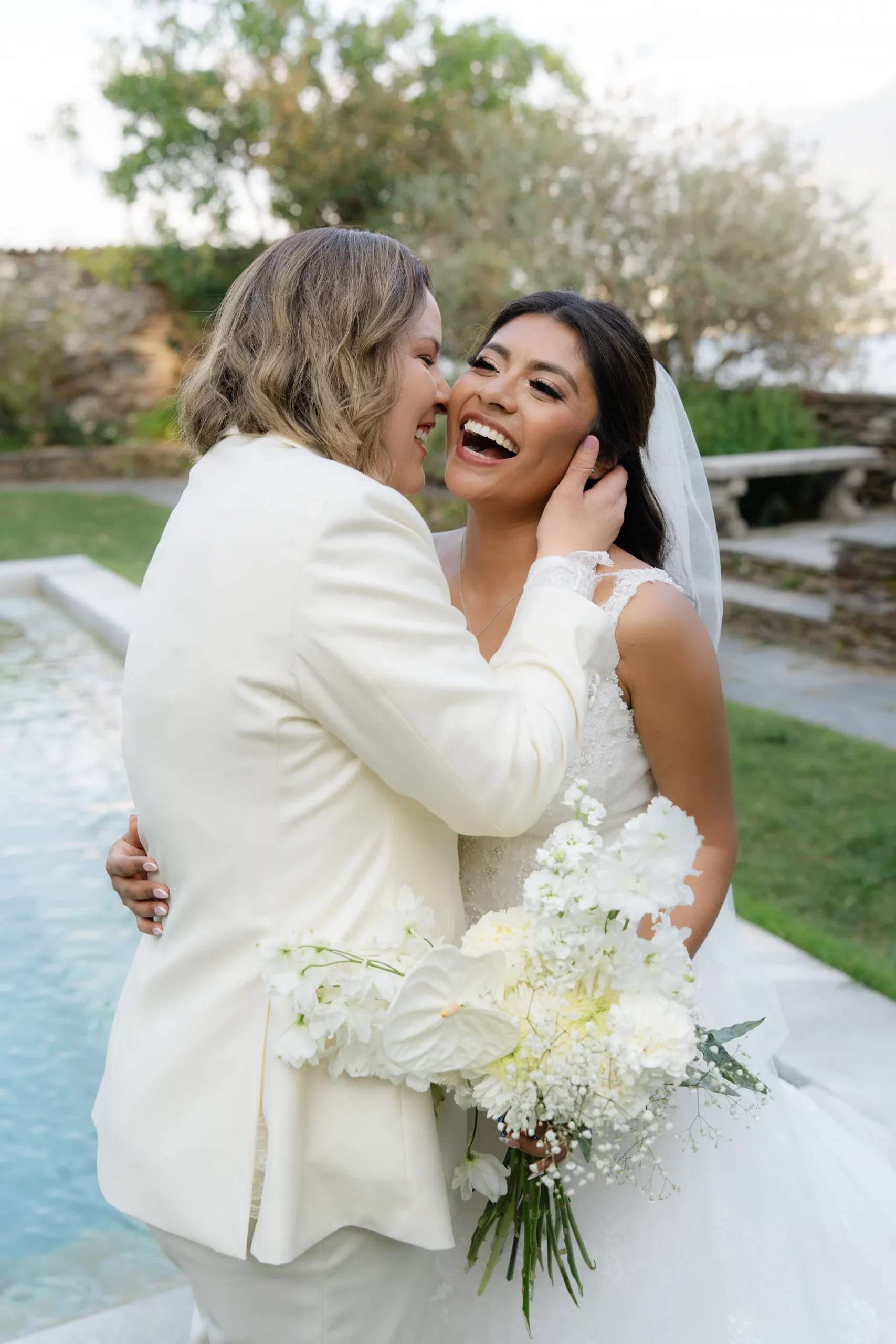 Ashley and Dionne laugh together poolside at Villa Emden on Brissago Island, Switzerland, holding a white bouquet.