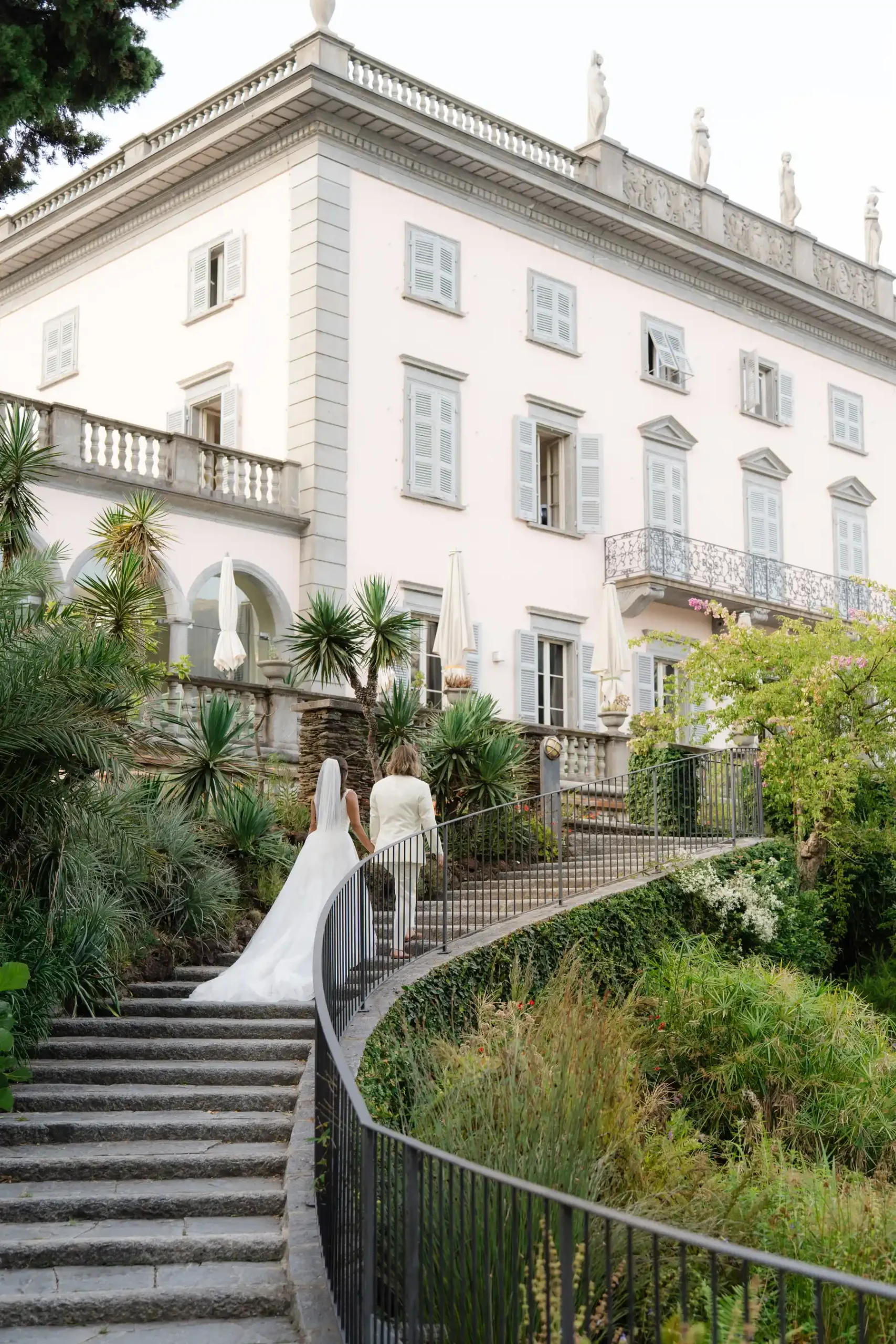 Ashley and Dionne walk up stone steps toward Villa Emden on Brissago Island, Switzerland, in wedding attire.