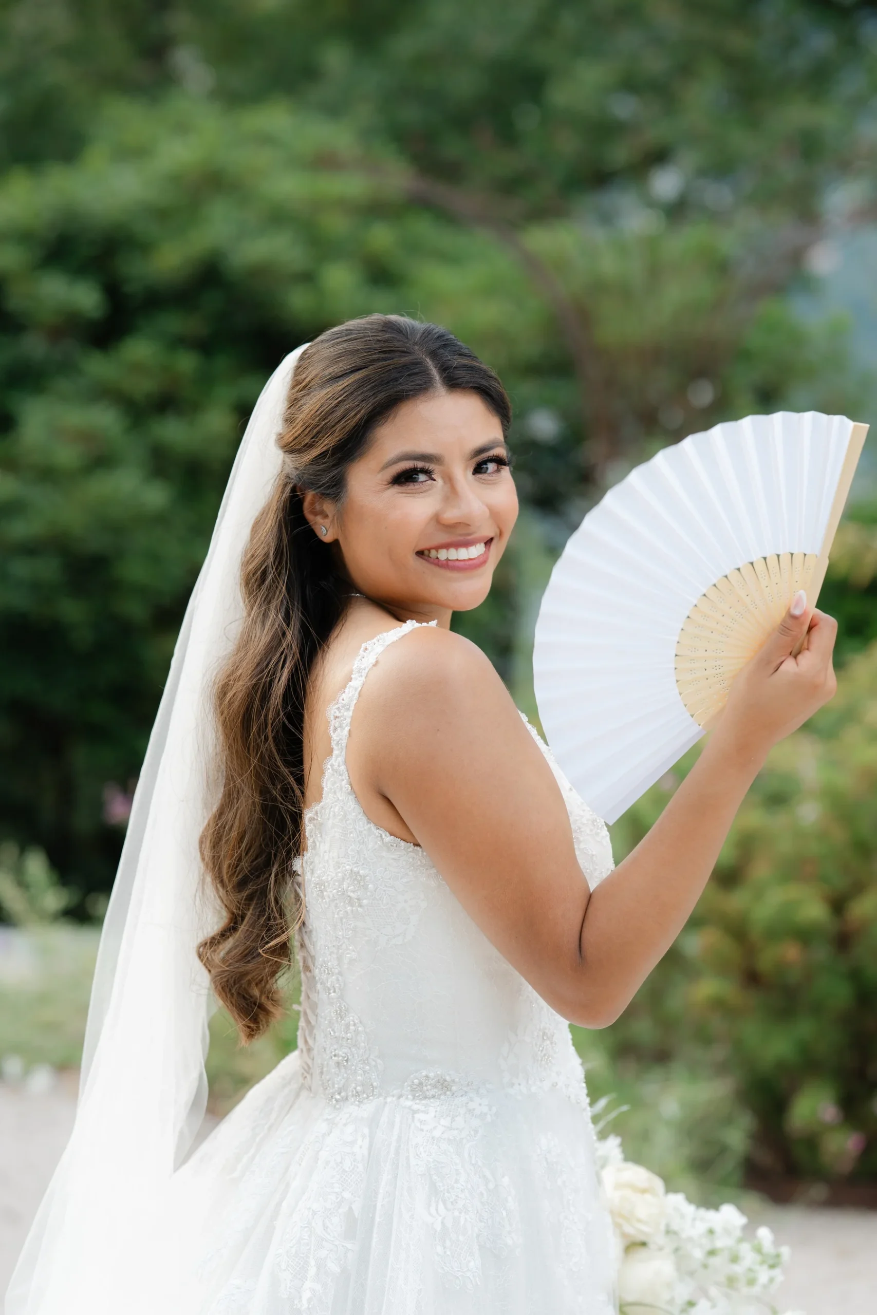 Bride Ashley Rodriguez-Vargas smiles over her shoulder in a lace gown and veil, holding a white fan at Villa Emden, Brissago Island, Switzerland.