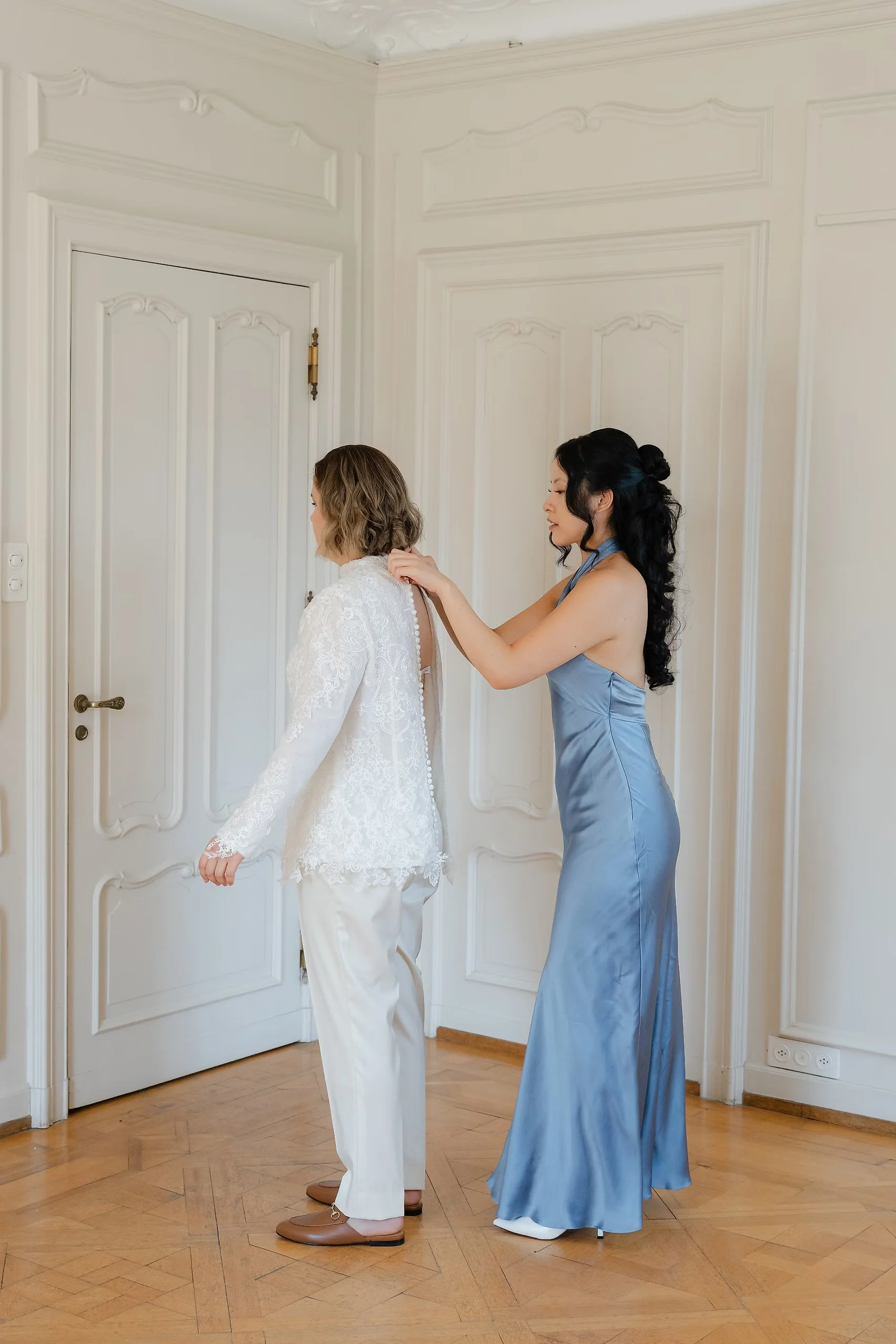 A bridesmaid in a blue satin gown buttons the back of a bride's lace bridal top at Villa Emden, Brissago Island.
