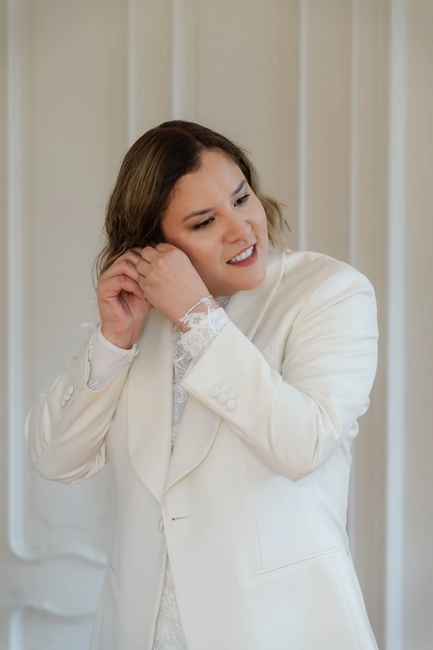 Bride smiles while putting on an earring, wearing a white blazer and lace top before her wedding at Villa Emden.