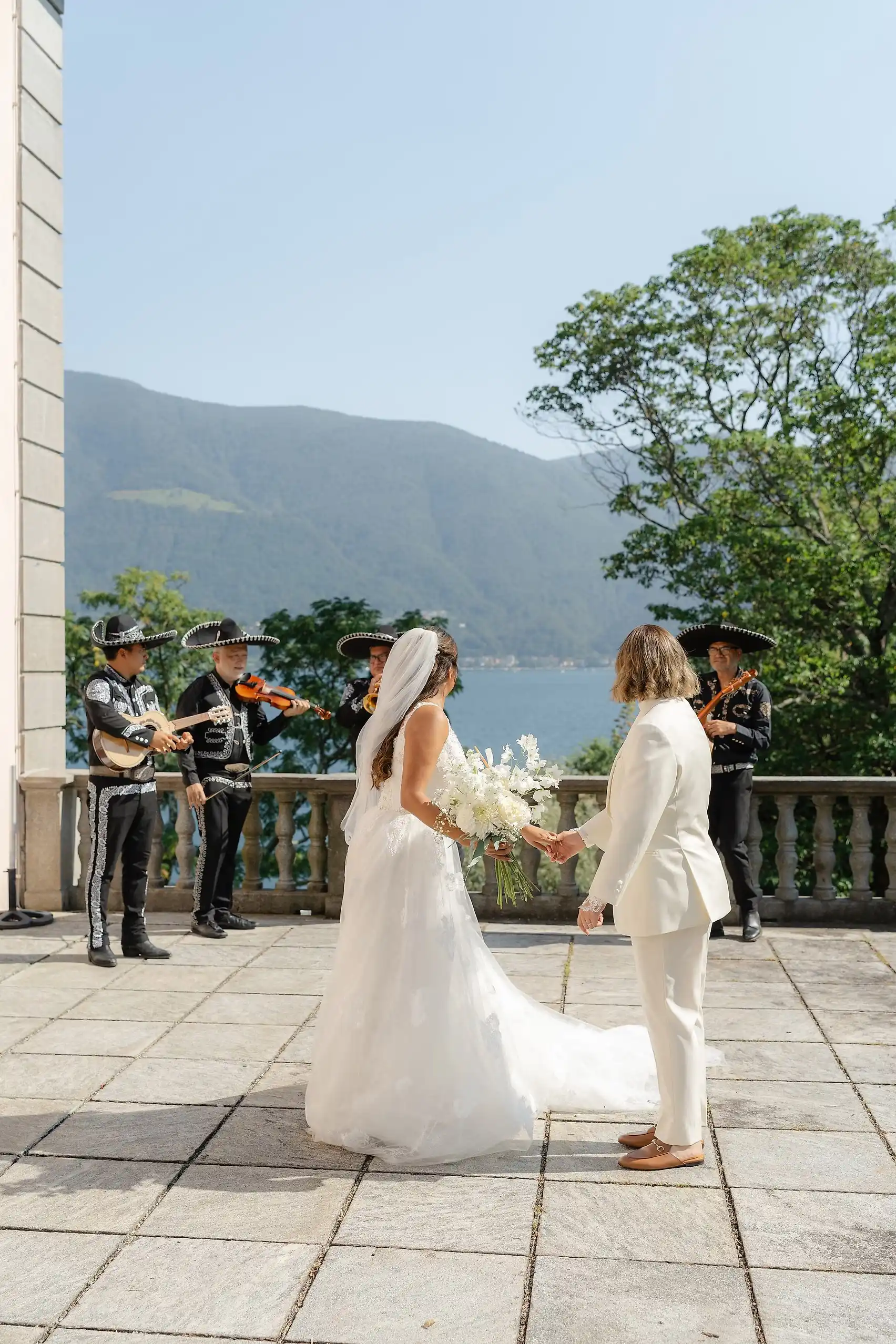 Ashley and Dionne hold hands on a terrace at Villa Emden, Brissago Island, Switzerland, as a mariachi band plays behind them.