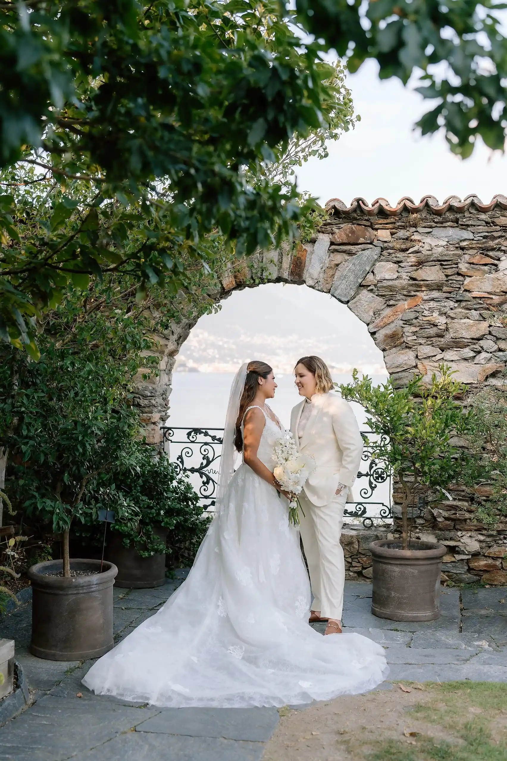 Ashley and Dionne Vargas stand face-to-face in wedding attire beneath a stone arch at Villa Emden on Brissago Island, Switzerland.
