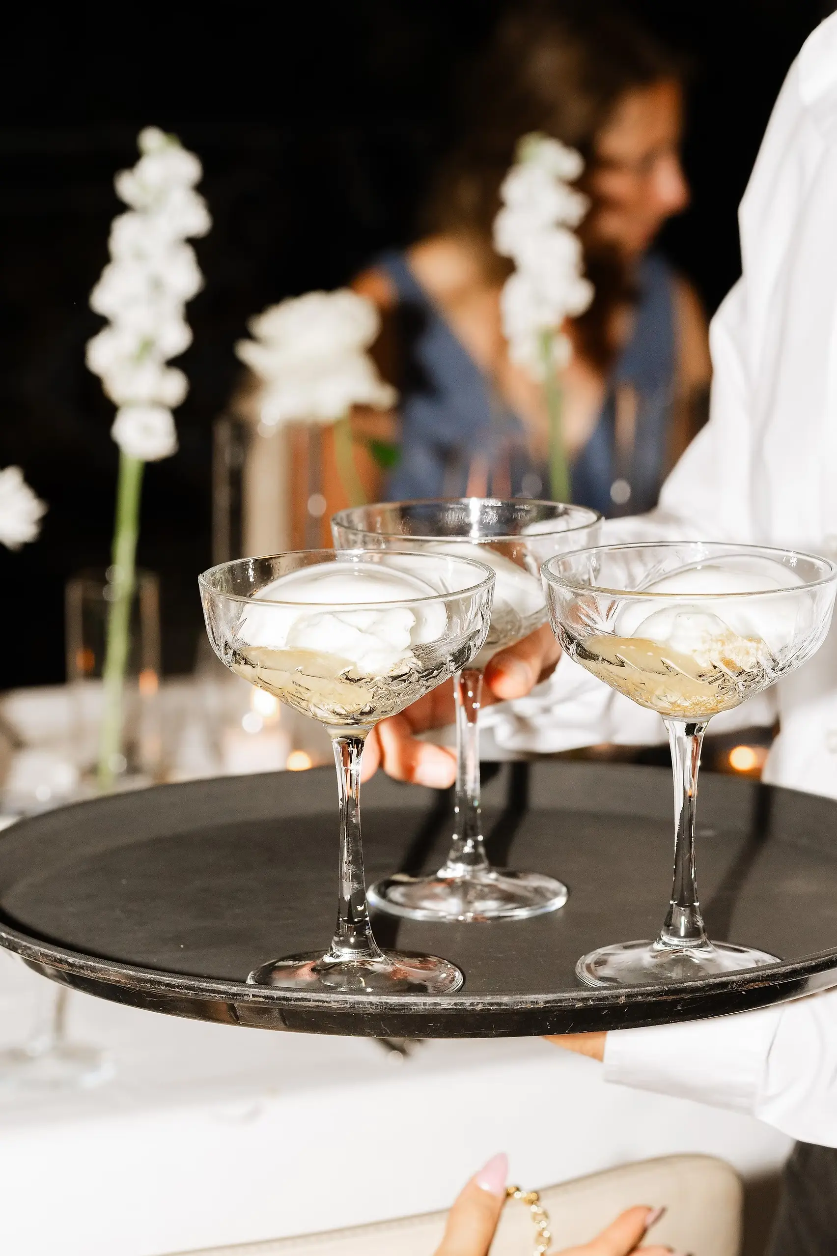 A server carries three crystal coupe glasses with white foam cocktails on a black tray at Villa Emden on Brissago Island.